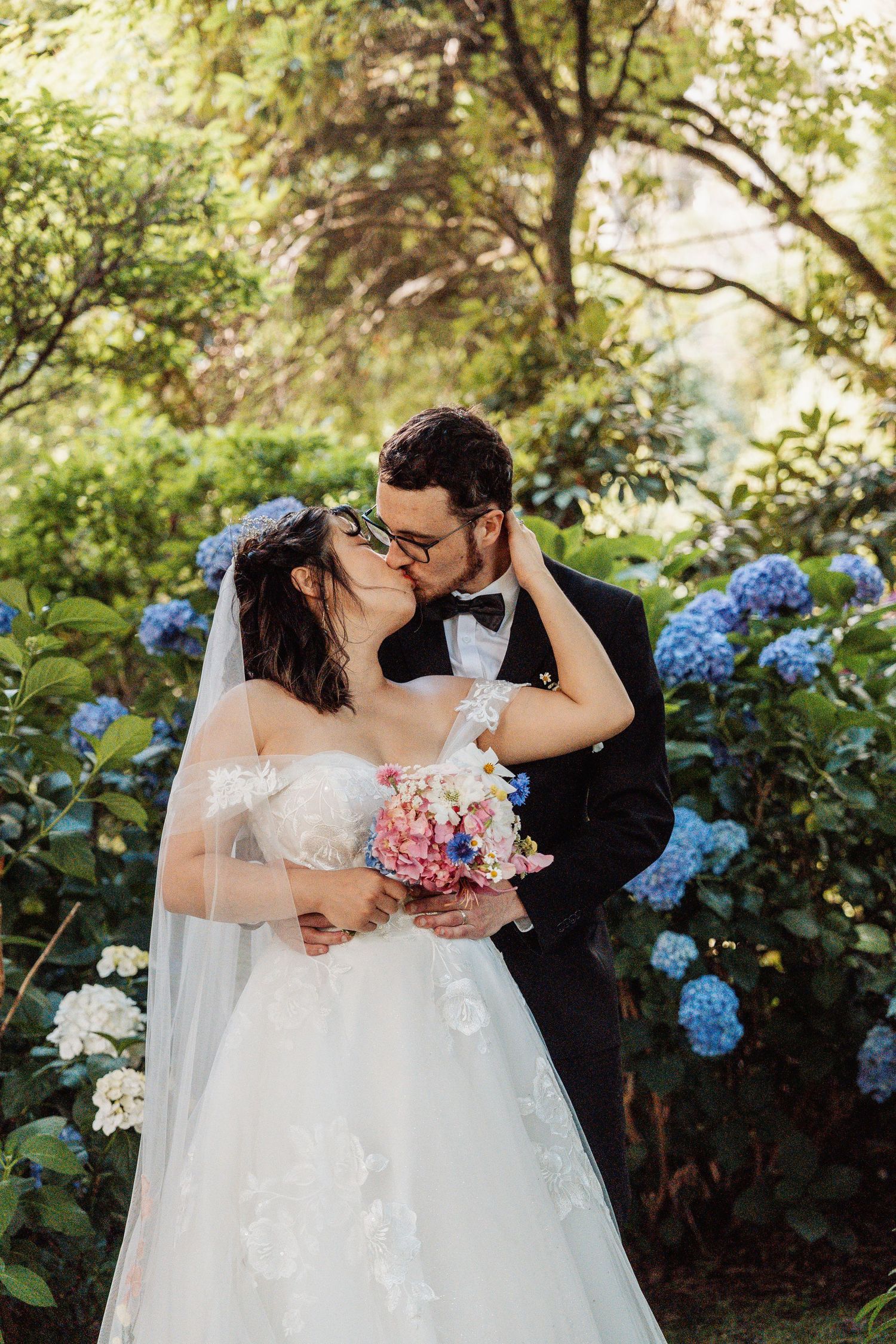 Bride and groom sharing a kiss in a lush garden with blue hydrangeas, bride holding a colorful bouquet.