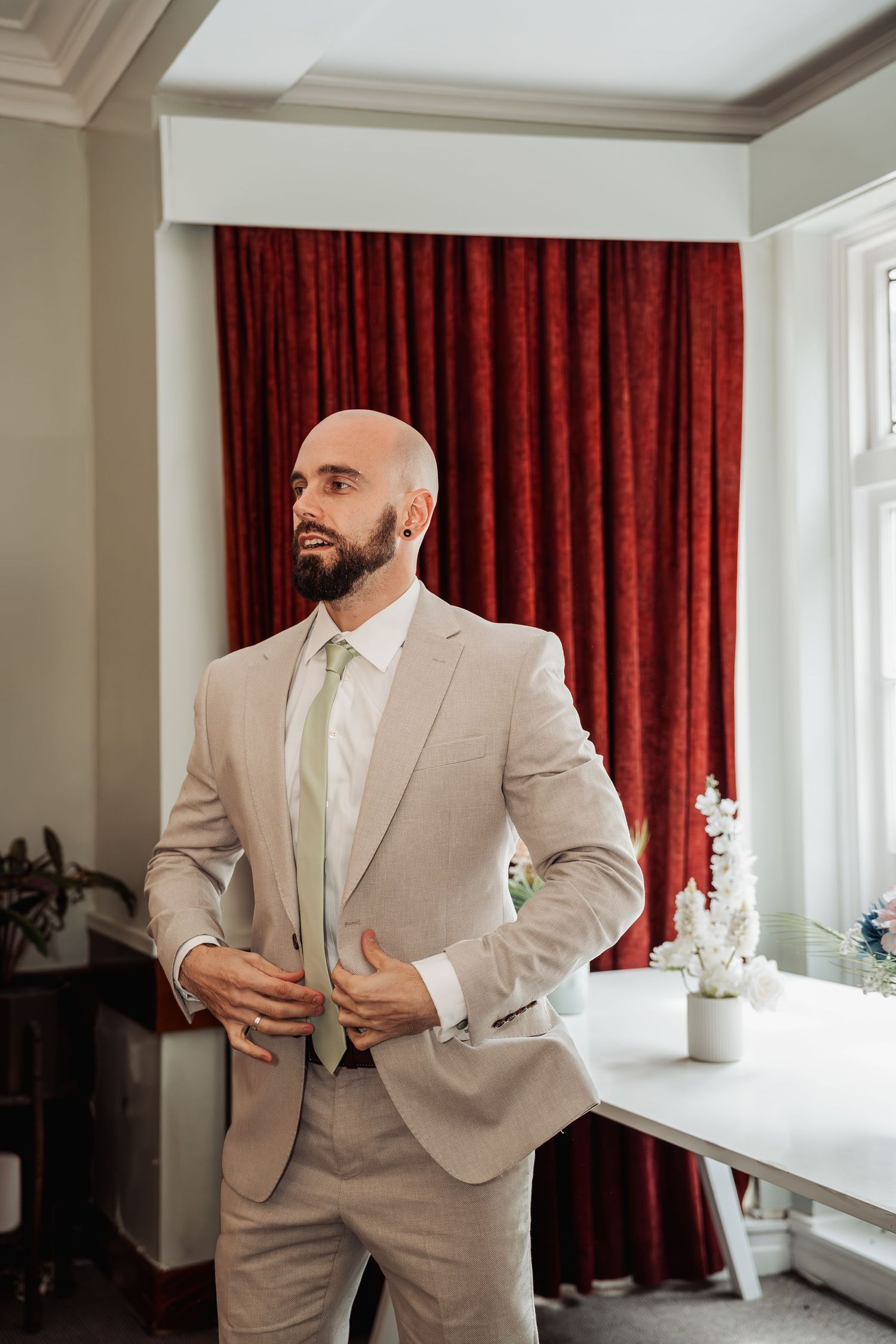 Bald groom with beard adjusting his beige suit jacket and green tie before wedding ceremony.