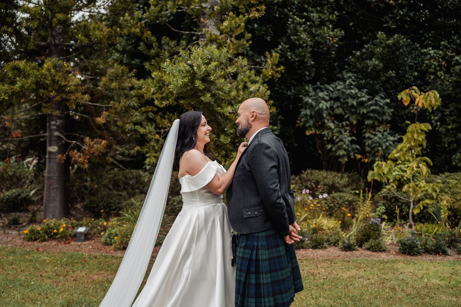 Bride in white off-shoulder gown and groom in tartan kilt hold hands in lush garden setting.