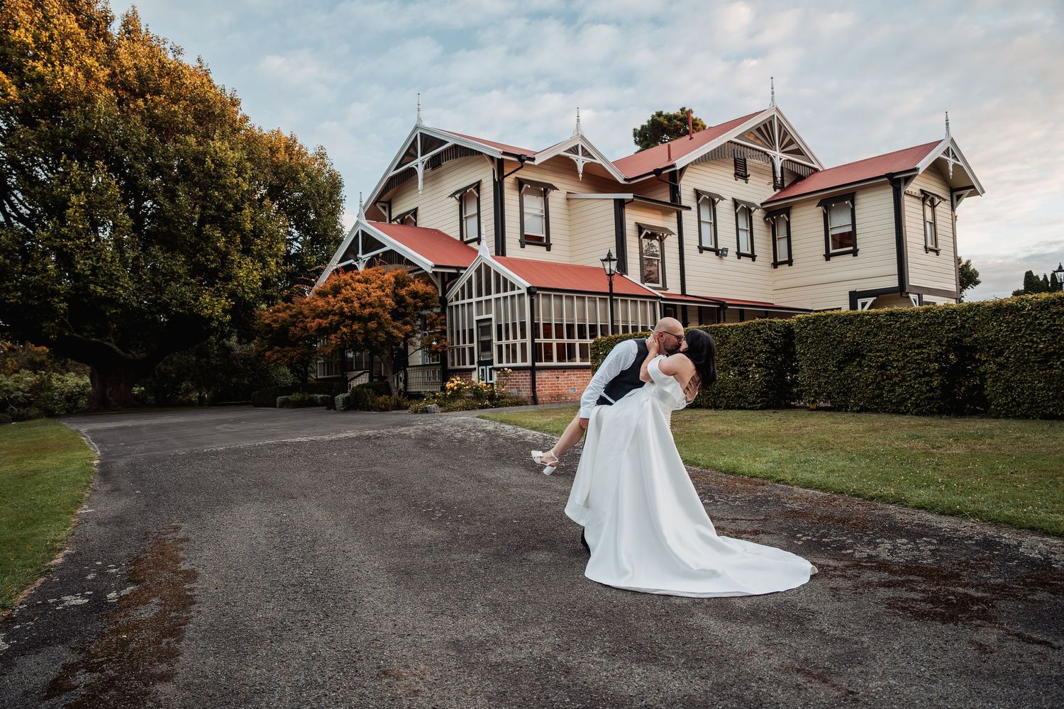 Groom dipping bride in white gown outside elegant historic manor house with red roof on wedding day.
