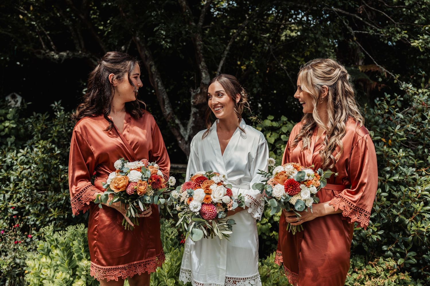 Bride in white robe with two bridesmaids in rust satin robes holding colorful bouquets outdoors.