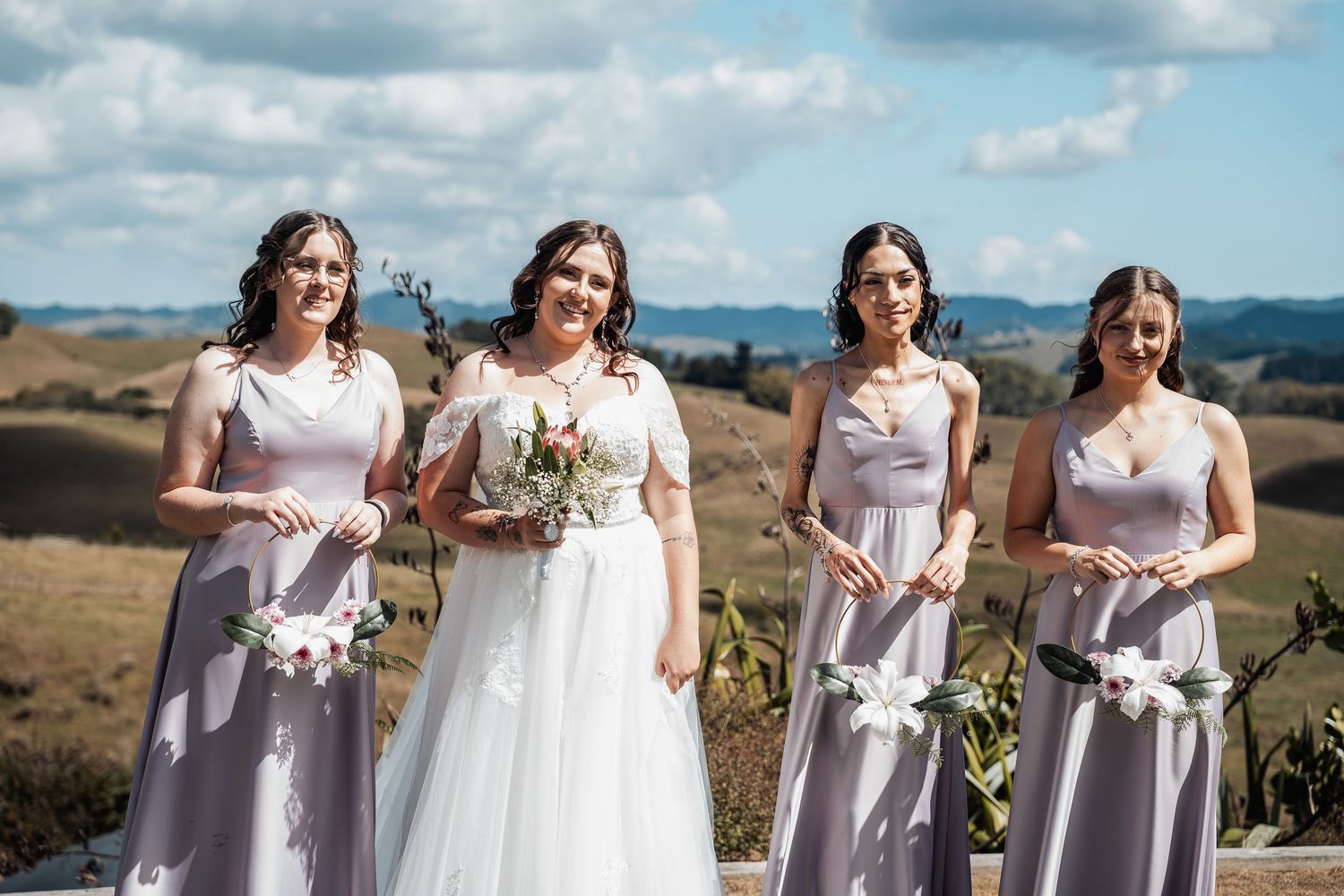 Bride in white gown holds bouquet with three bridesmaids in lavender dresses against rolling hills backdrop.