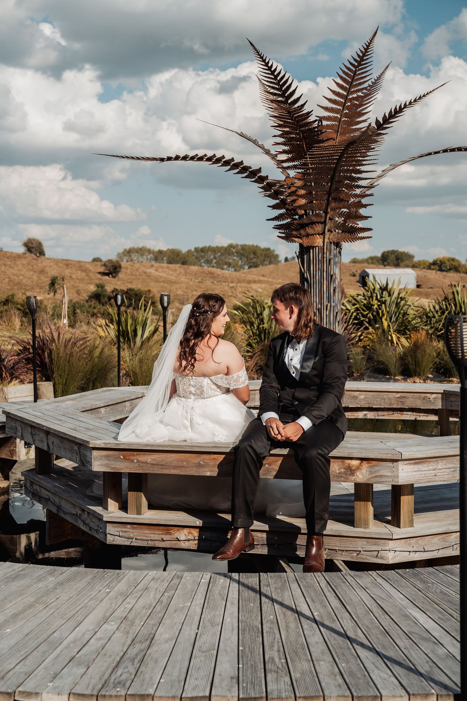Newlywed couple sits together on wooden deck gazing at each other near decorative bronze fern sculpture.