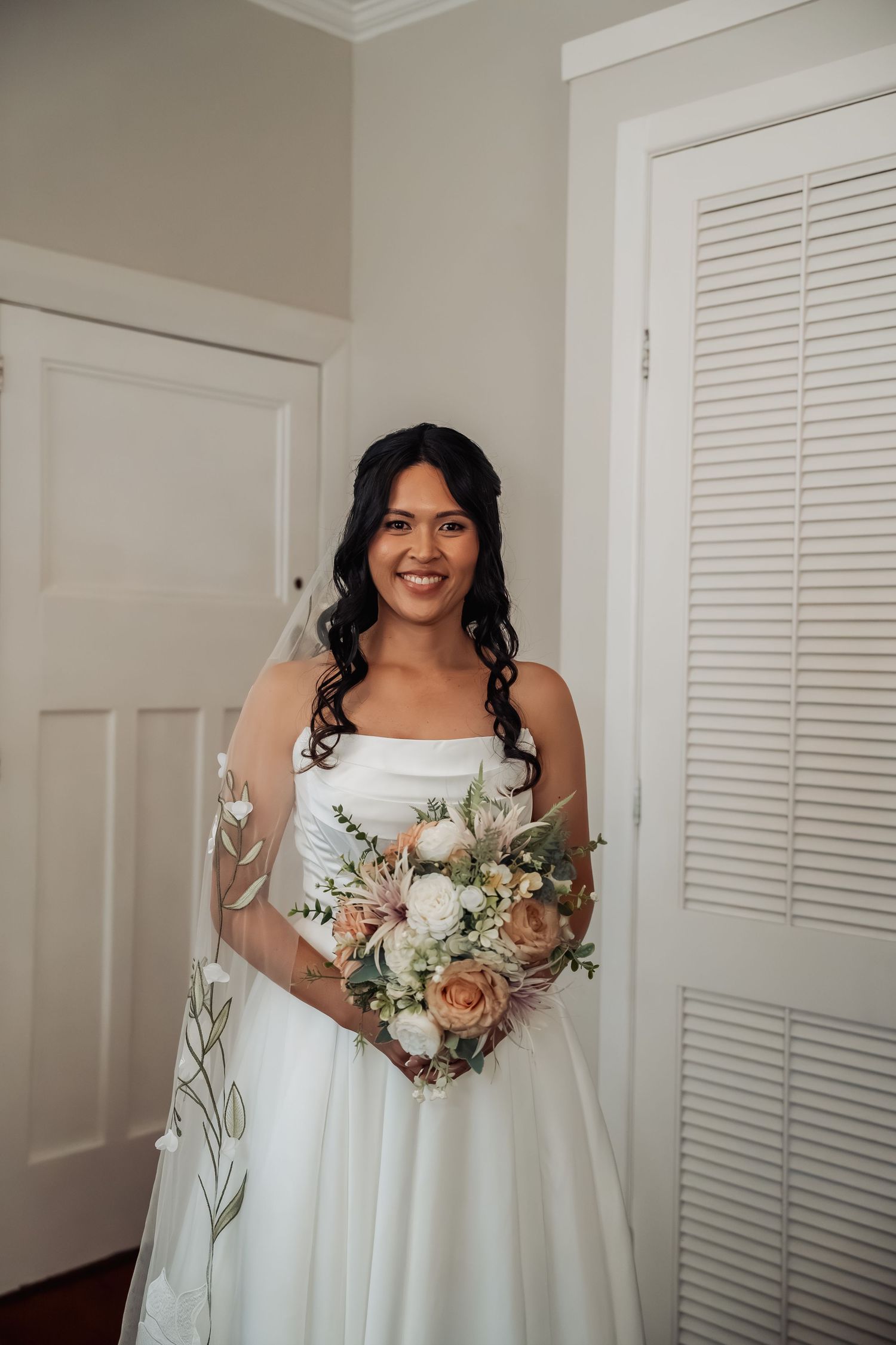 Smiling bride in strapless white wedding dress holds bouquet of blush and white roses with eucalyptus.