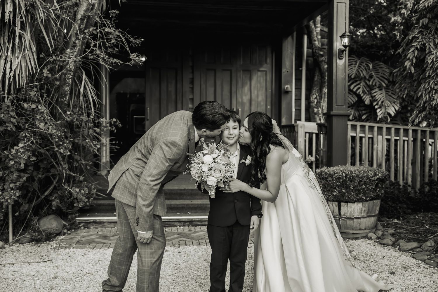 Black and white photo of bride and groom leaning in to kiss young boy holding bridal bouquet.