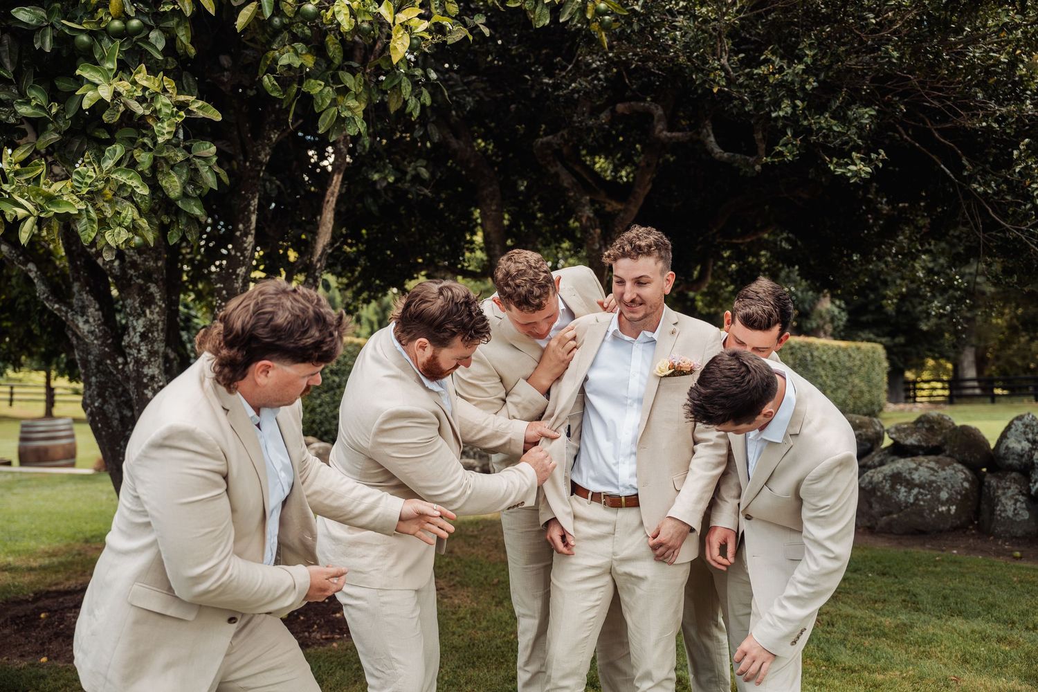 Groomsmen in beige suits sharing a fun candid moment together in a garden setting.