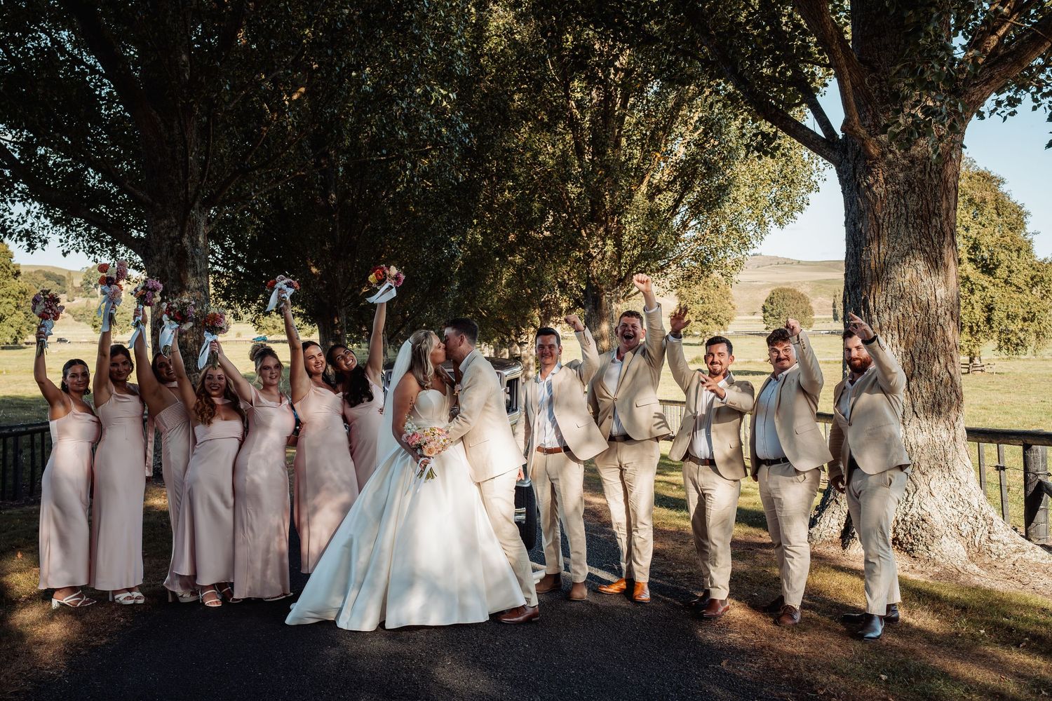 Wedding party in beige suits and blush gowns celebrate joyfully under large trees on a sunny wedding day.
