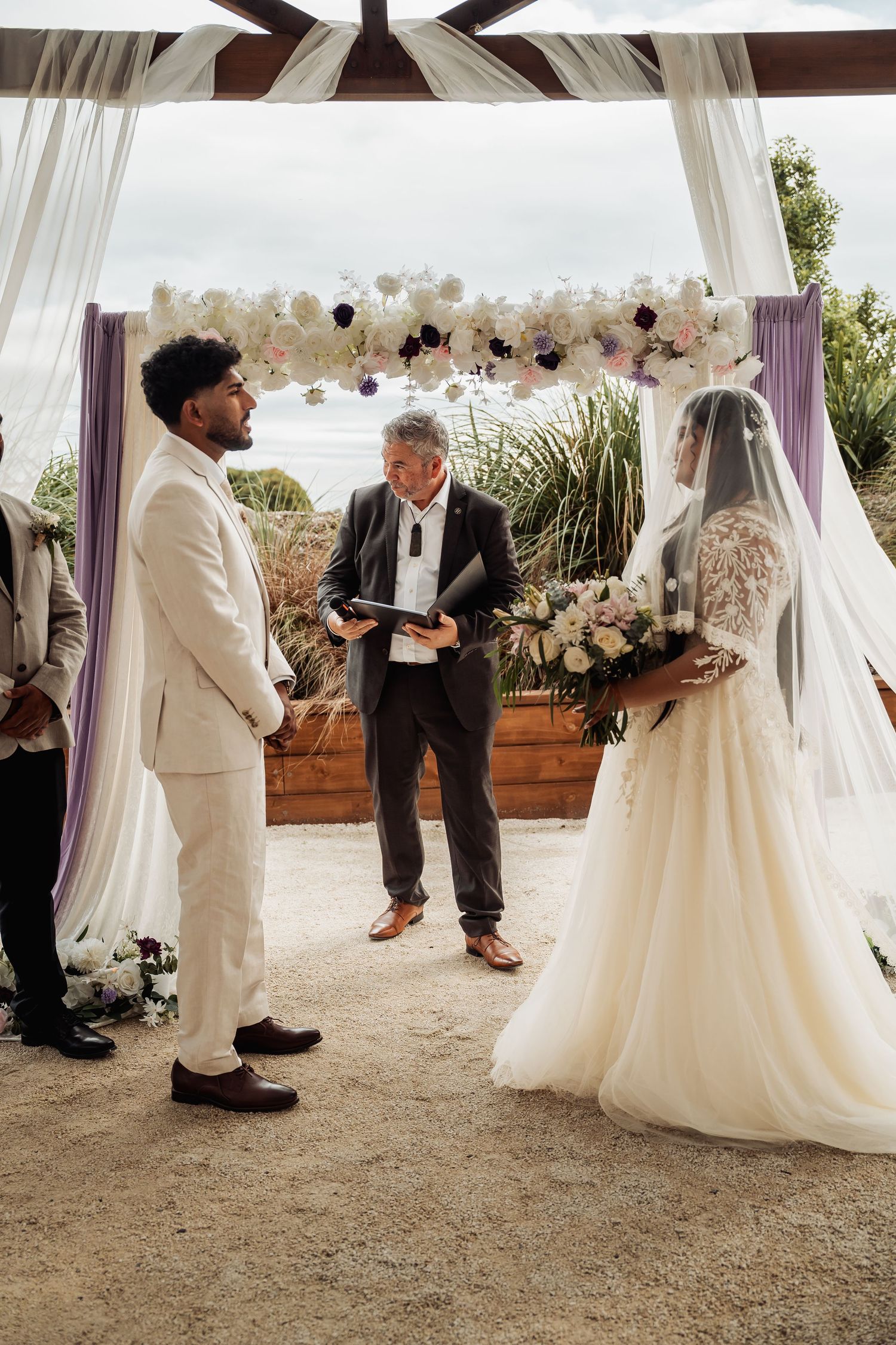 Couple exchanges vows under floral arch with officiant at outdoor seaside wedding ceremony.