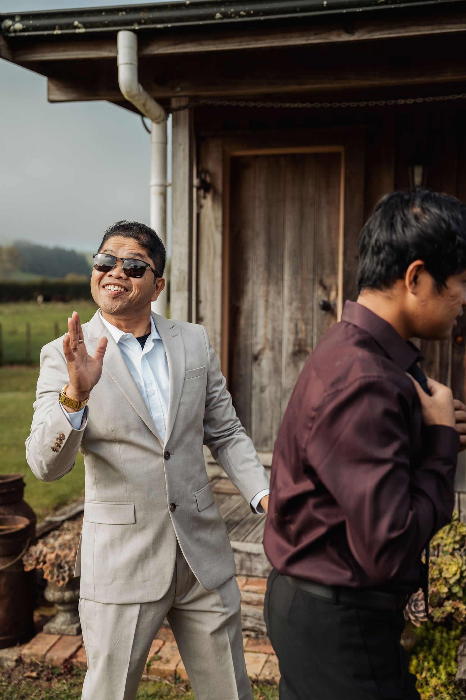 Groom in gray suit and sunglasses waves and smiles outside rustic barn at wedding.