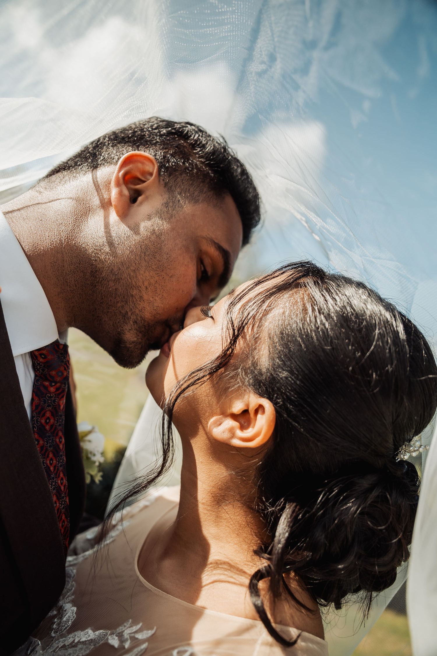 Romantic close-up of groom kissing bride on forehead under flowing white veil in natural sunlight.
