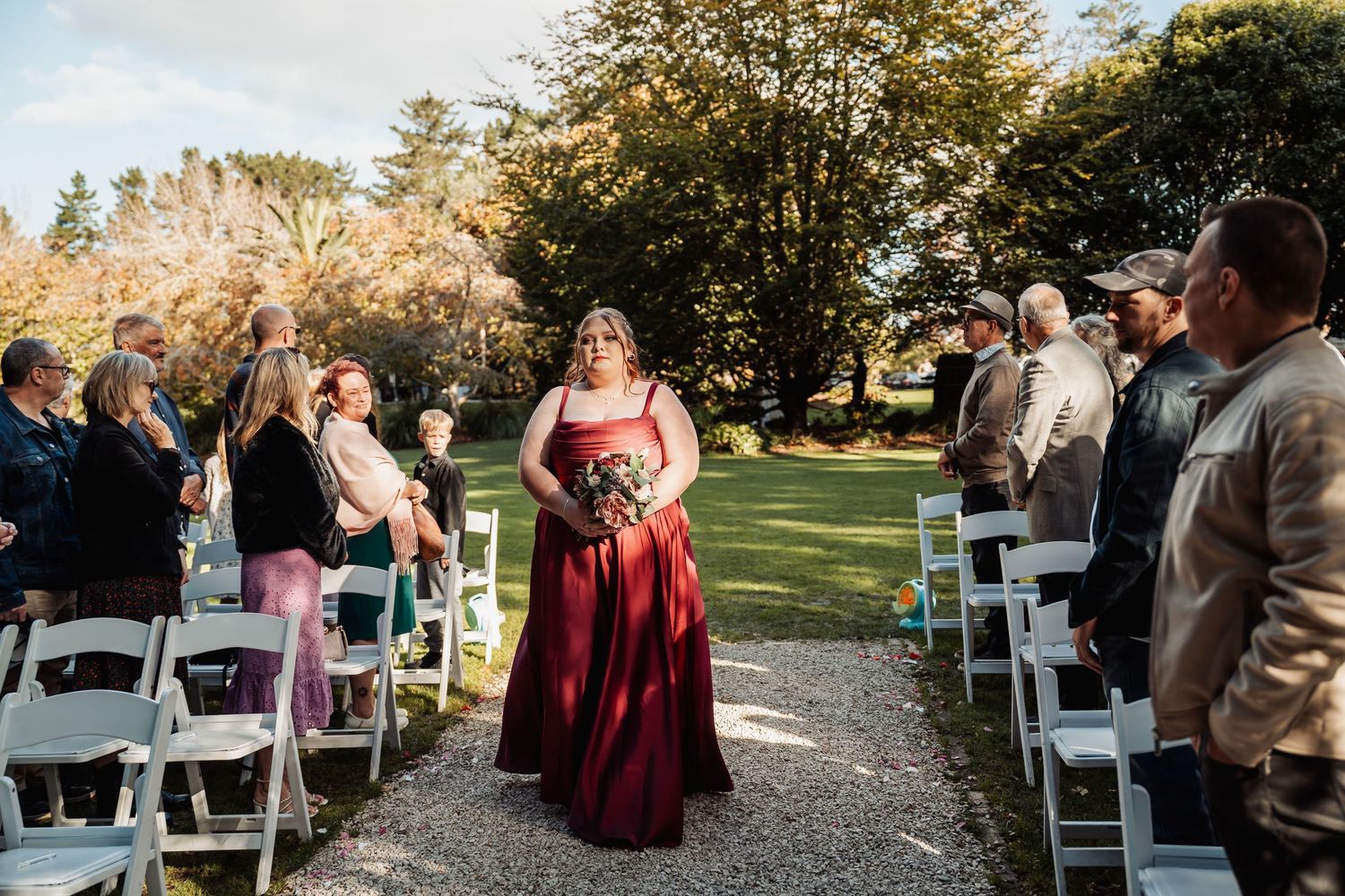 Bridesmaid in red gown walks down outdoor wedding aisle lined with white chairs and guests.