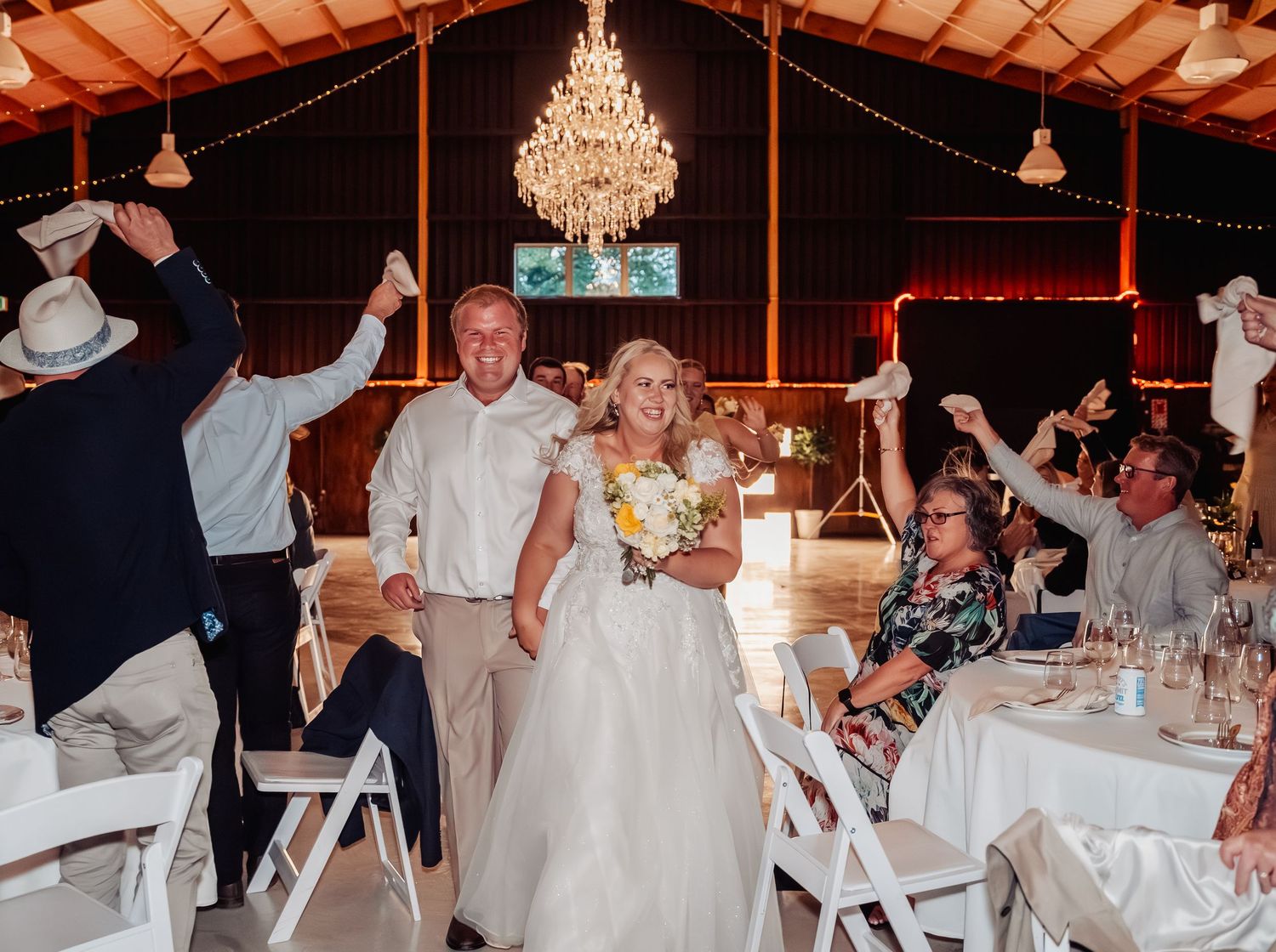 Bride and groom walk through cheering guests at rustic barn wedding reception under sparkling chandelier.