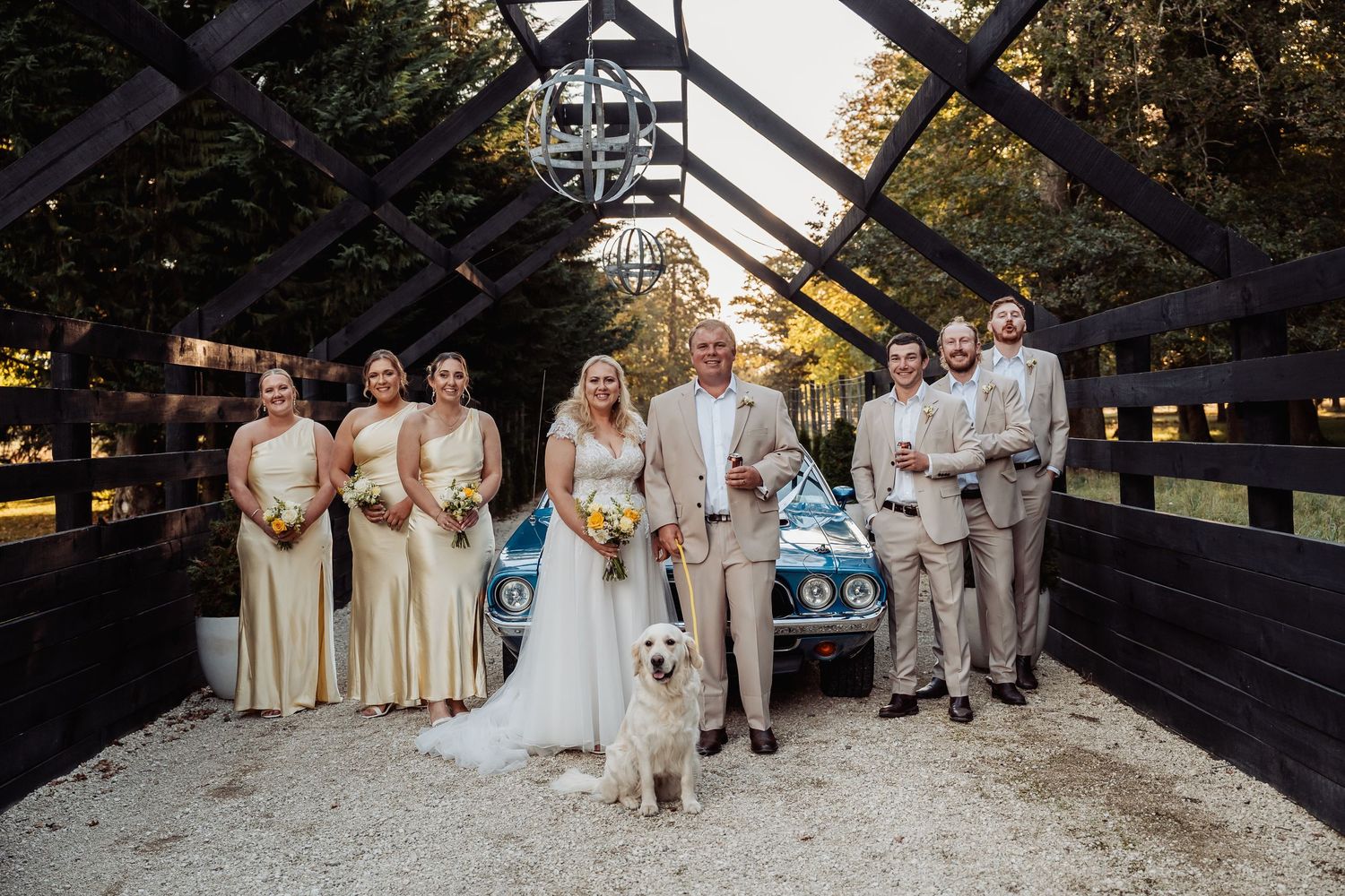 Wedding party with golden retriever and vintage blue car under rustic barn arch.