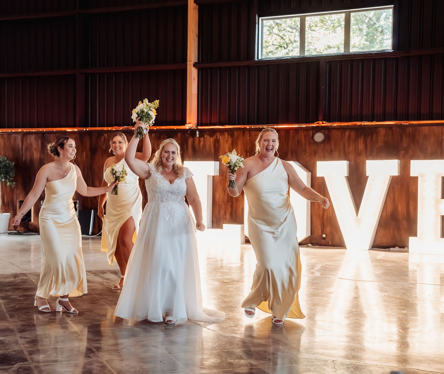 Bride and bridesmaids in ivory gowns celebrating in a rustic barn venue with large illuminated LOVE letters.