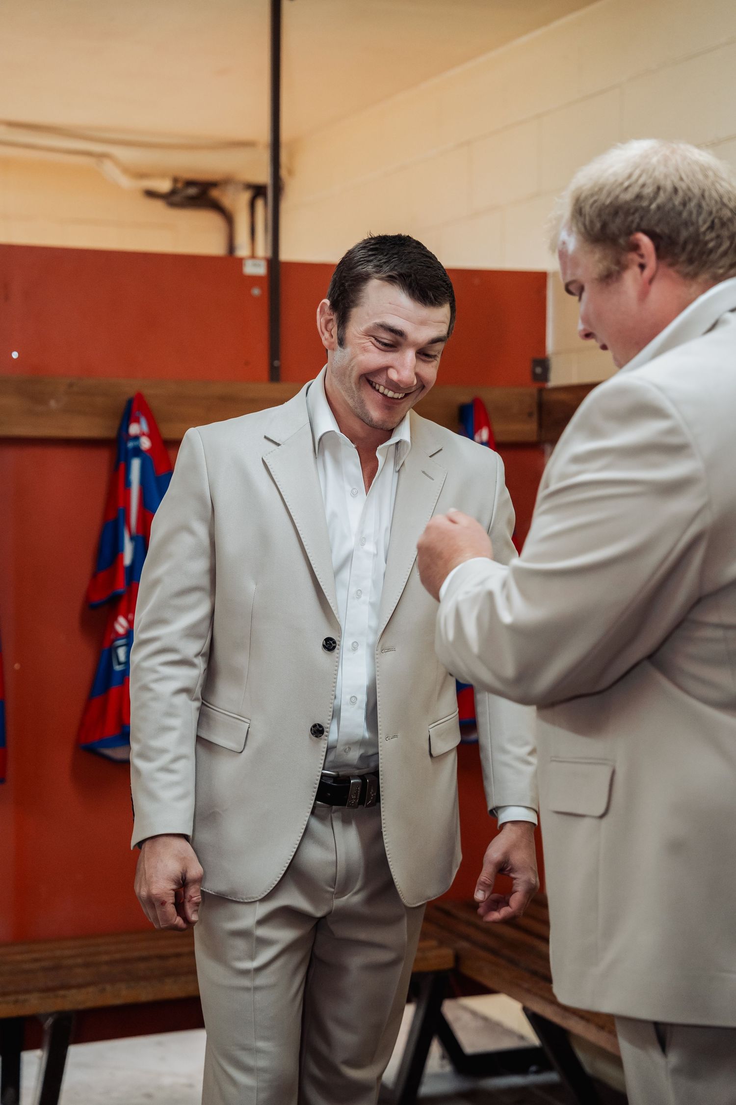 Two groomsmen in beige suits share a laugh while helping each other prepare in a locker room before a wedding.
