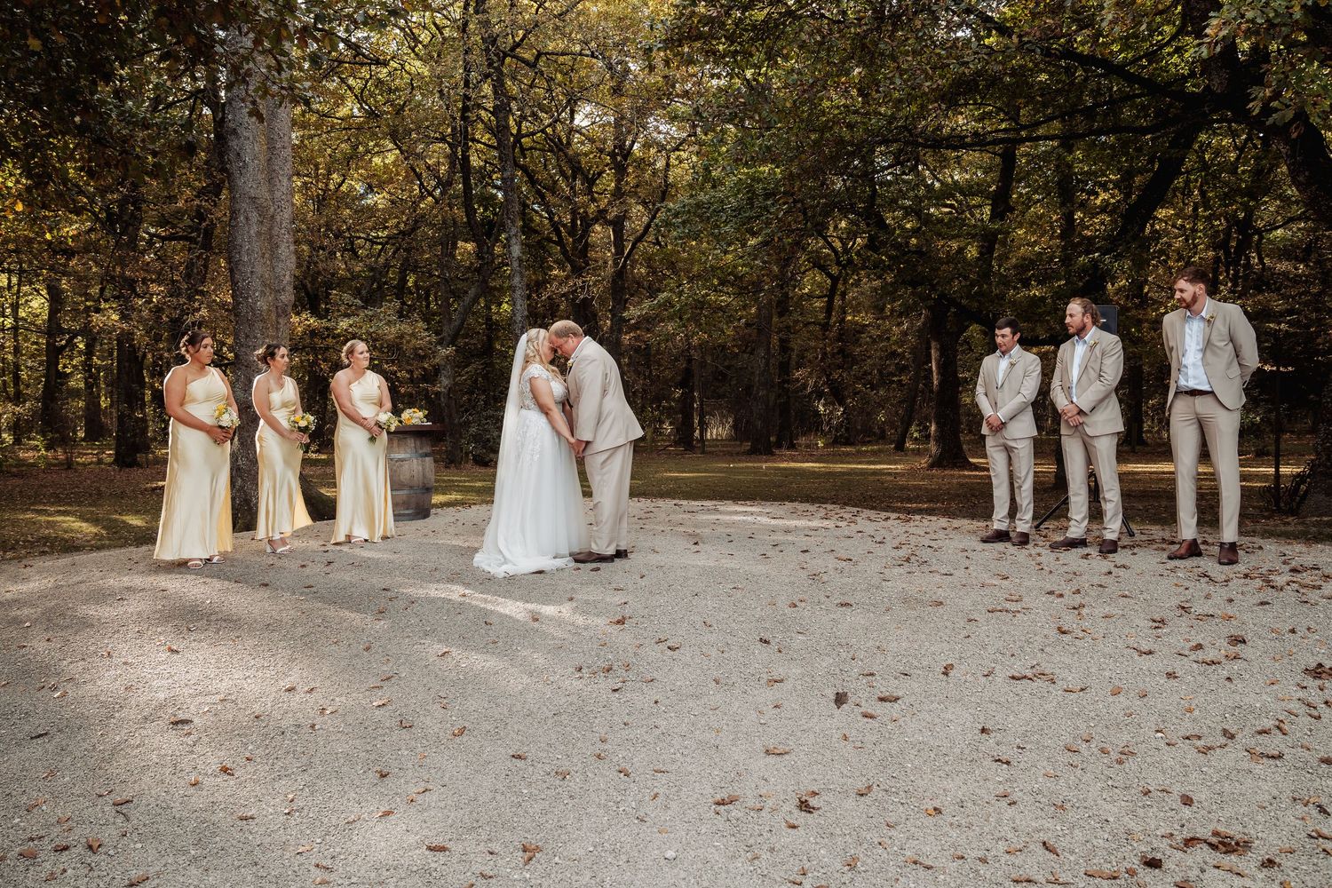 Wedding party stands on gravel path in wooded setting as bride and groom share first kiss.