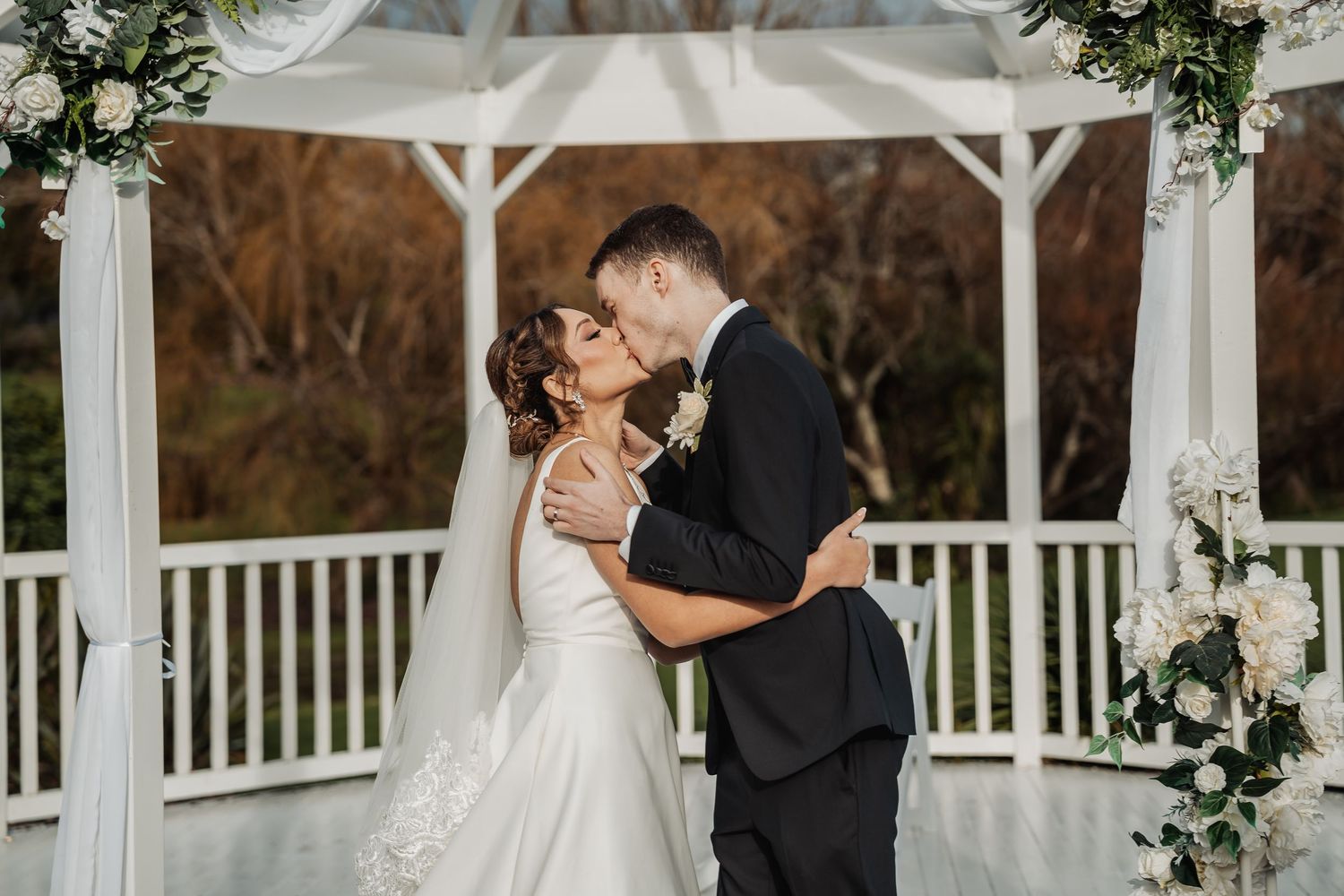 A romantic couple shares a kiss during their wedding ceremony at a white gazebo decorated with greenery.