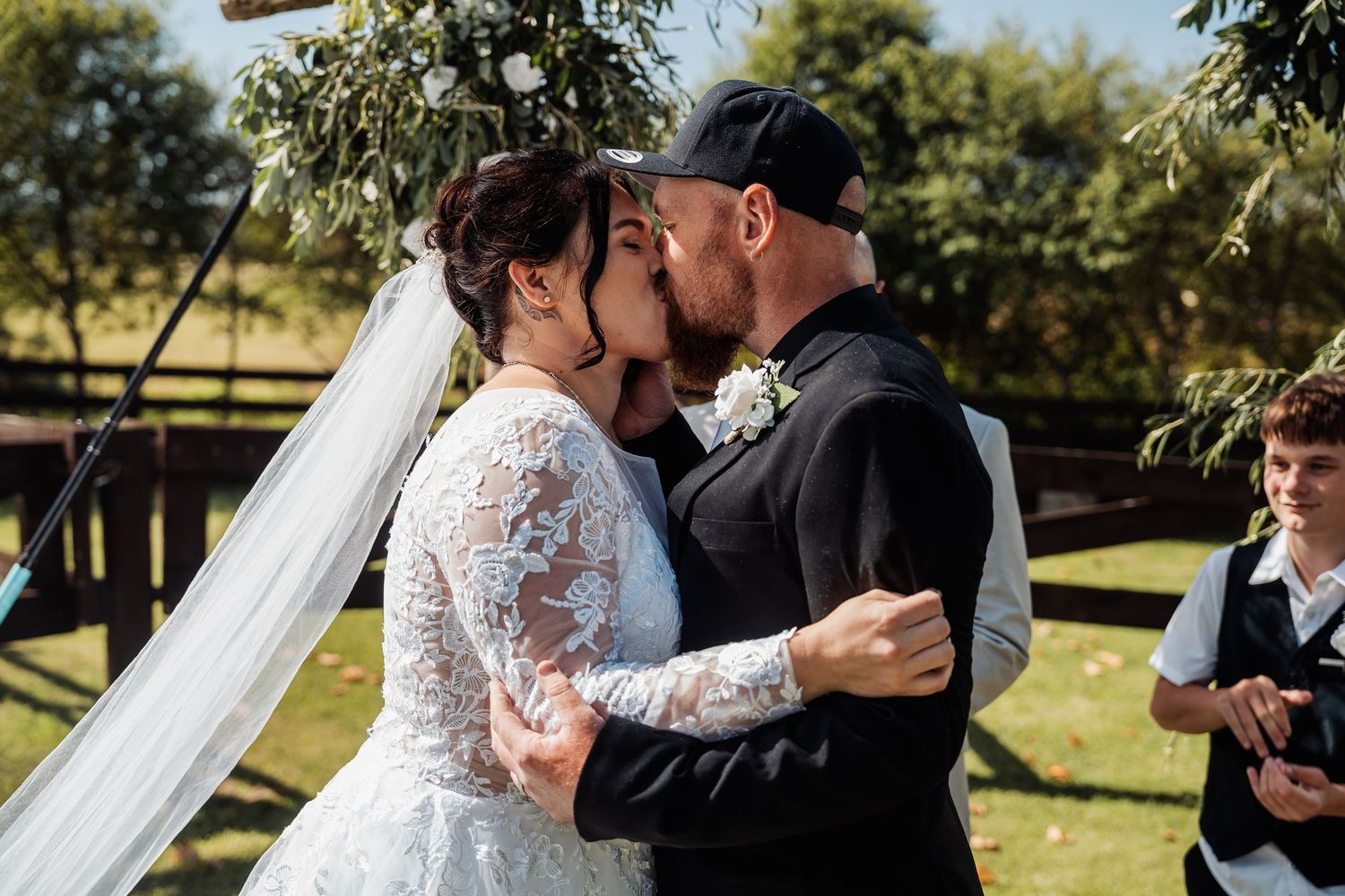 A couple shares their first kiss as newlyweds during an outdoor wedding ceremony on a sunny day.