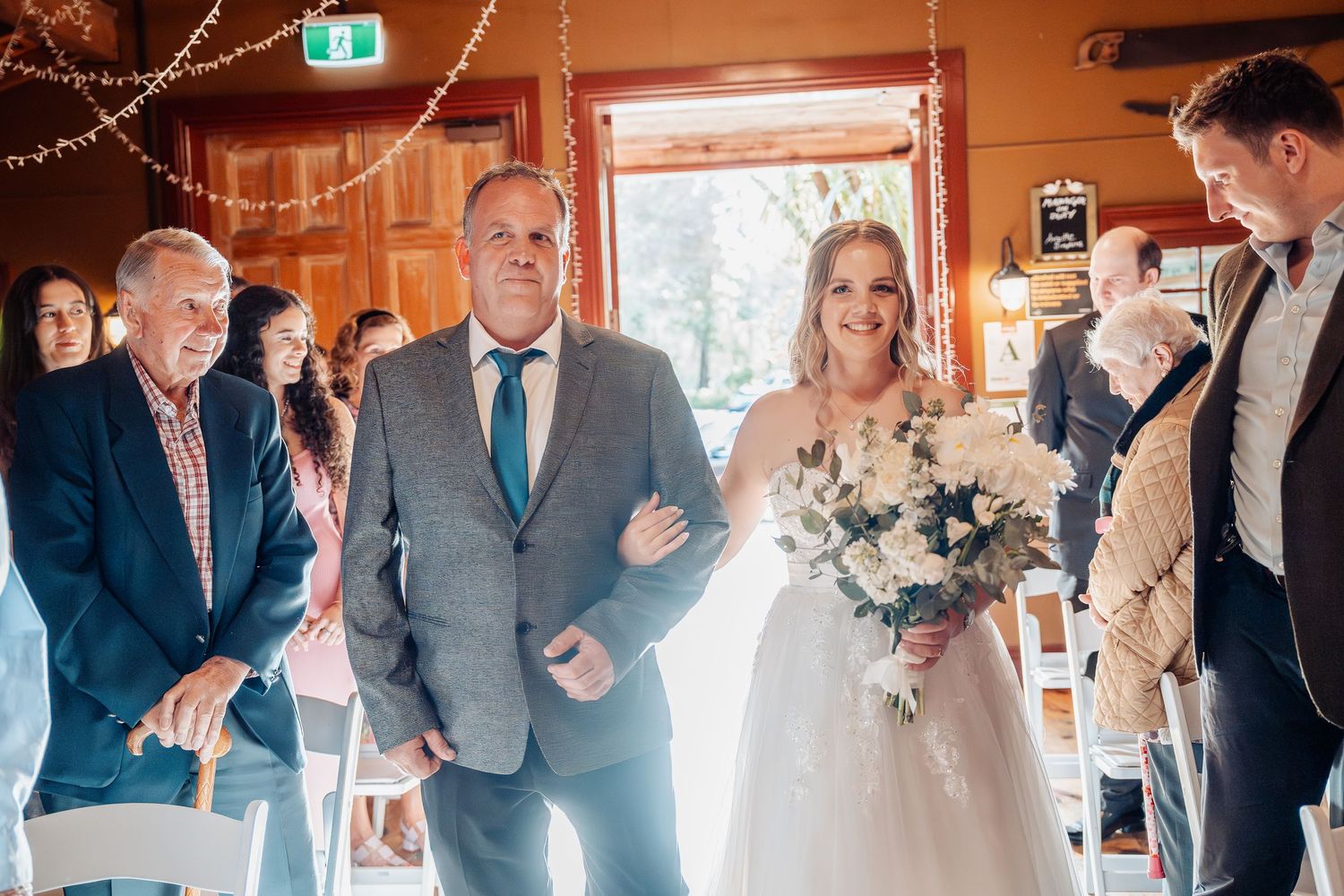 A wedding processional walking down an aisle in a rustic wooden interior with string lights overhead.