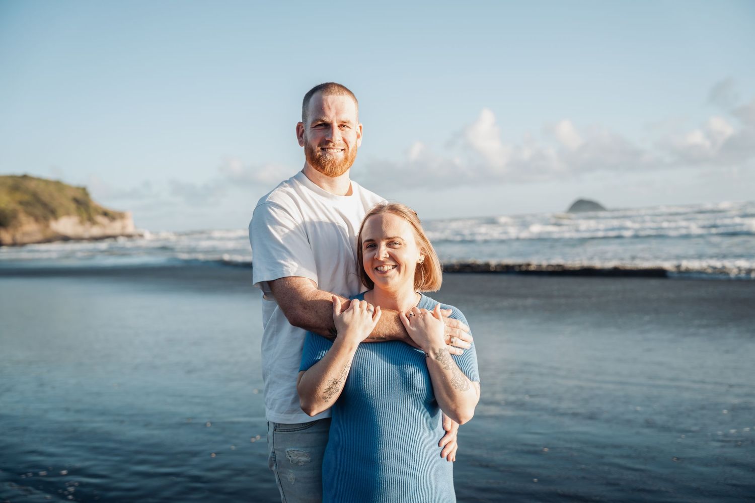 A loving couple poses on a scenic beach with waves and cliffs in the background during golden hour.