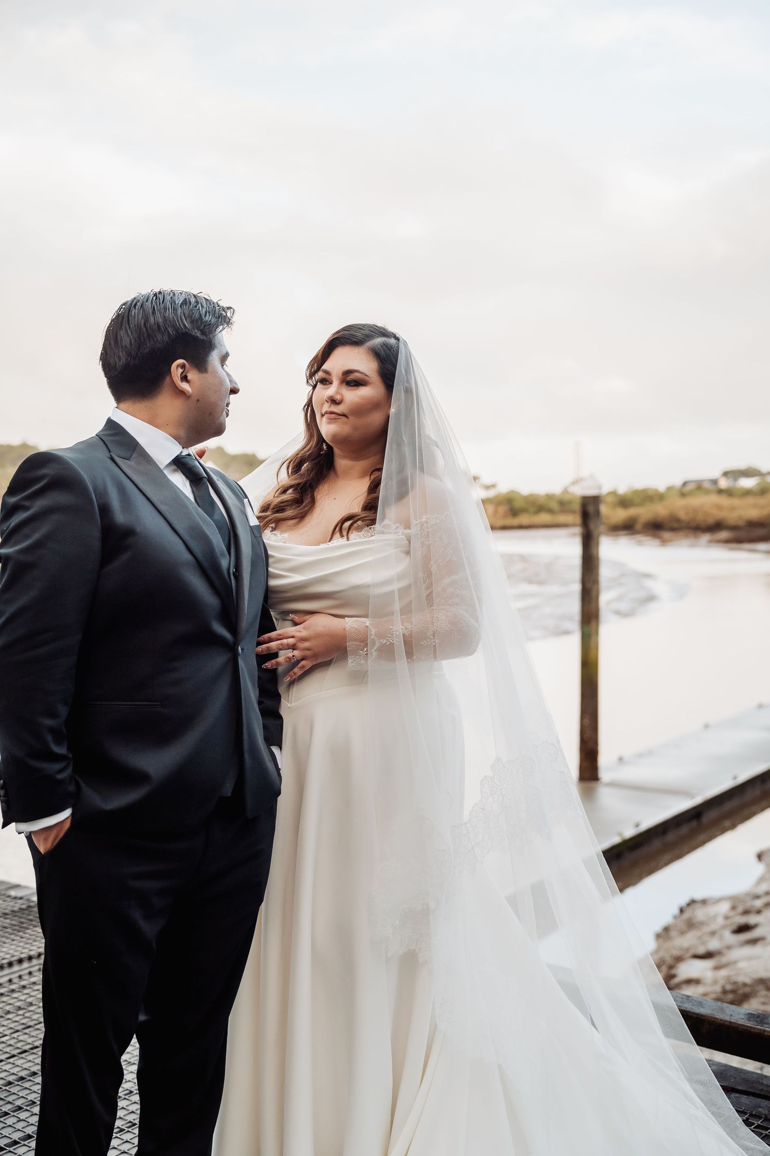 A wedding couple stands on a dock by the water, with the bride wearing a flowing white gown and veil.
