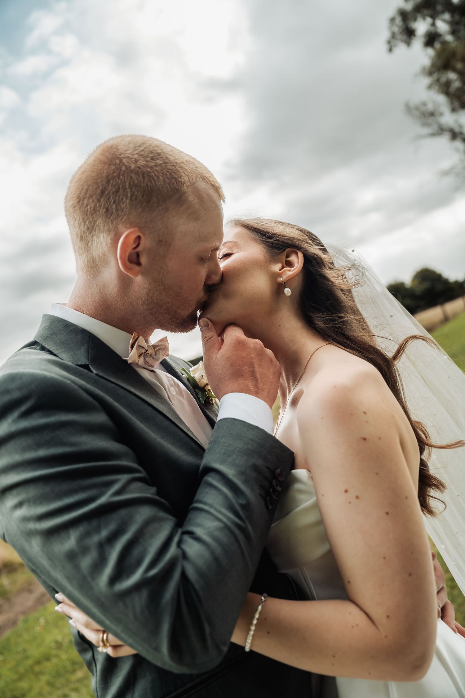 Groom tenderly cups bride's face during a romantic kiss outdoors on their wedding day under a cloudy sky.