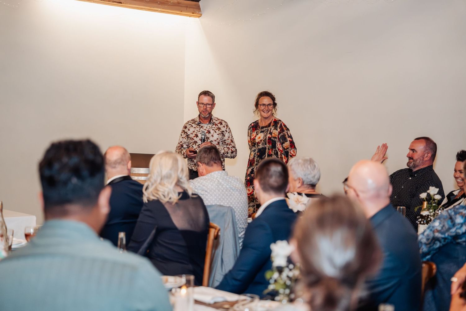 Two guests delivering a speech to seated wedding reception guests in a bright white venue.