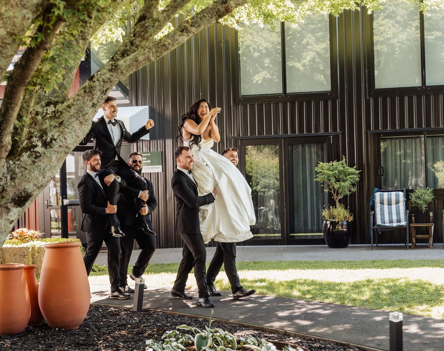 Groomsmen lift bride in white wedding dress outdoors near modern building surrounded by lush greenery.