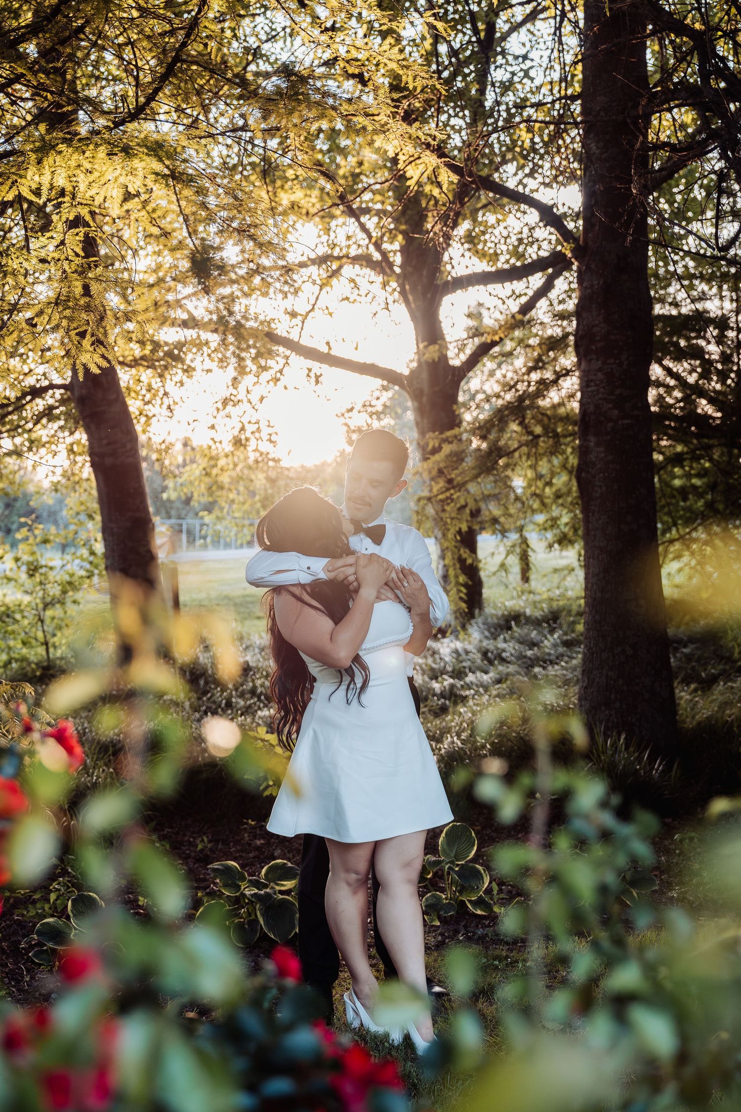 Couple embraces tenderly beneath golden sunlit trees in a romantic garden at sunset.