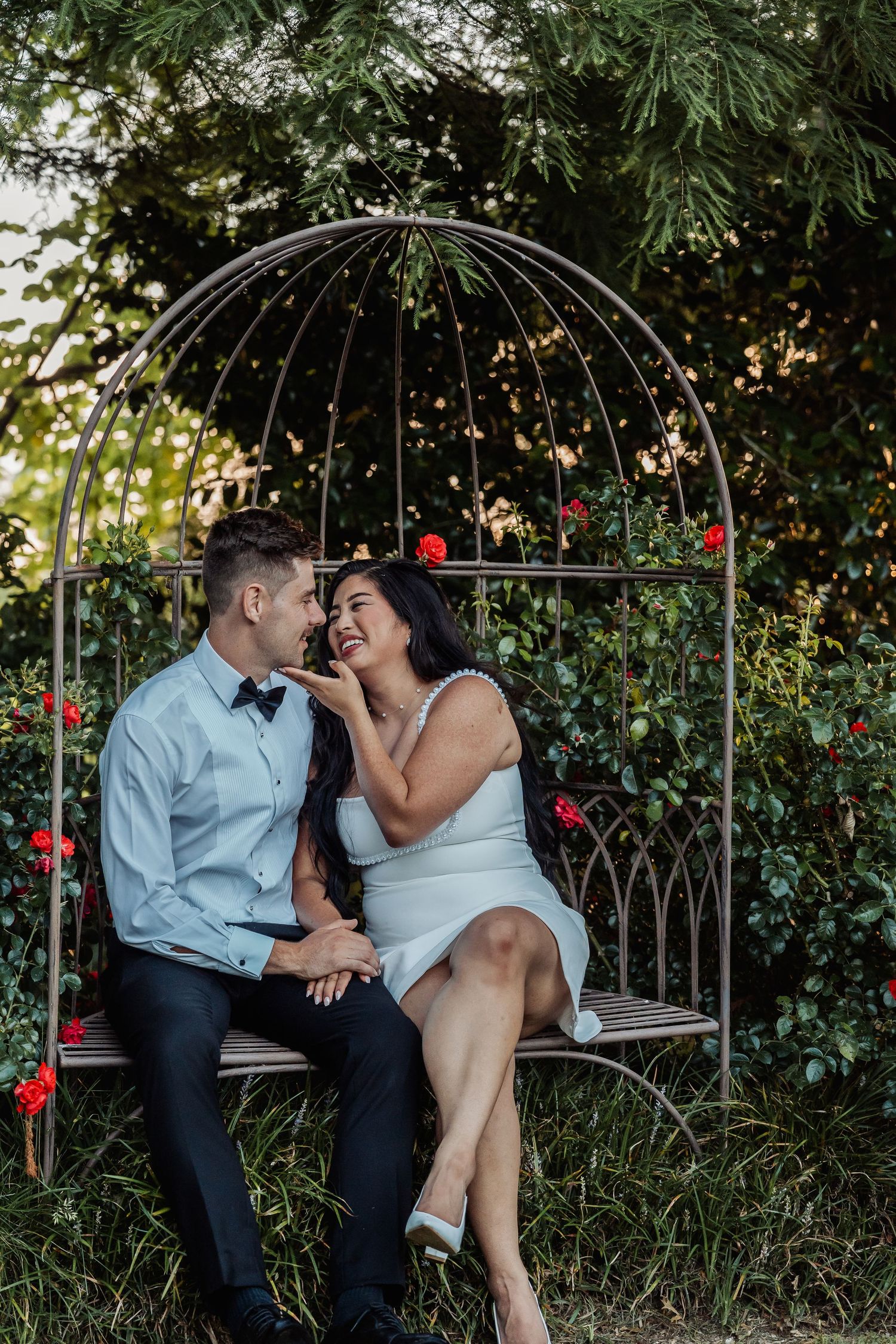 A couple laughs together while sitting on an ornate bench surrounded by red roses.