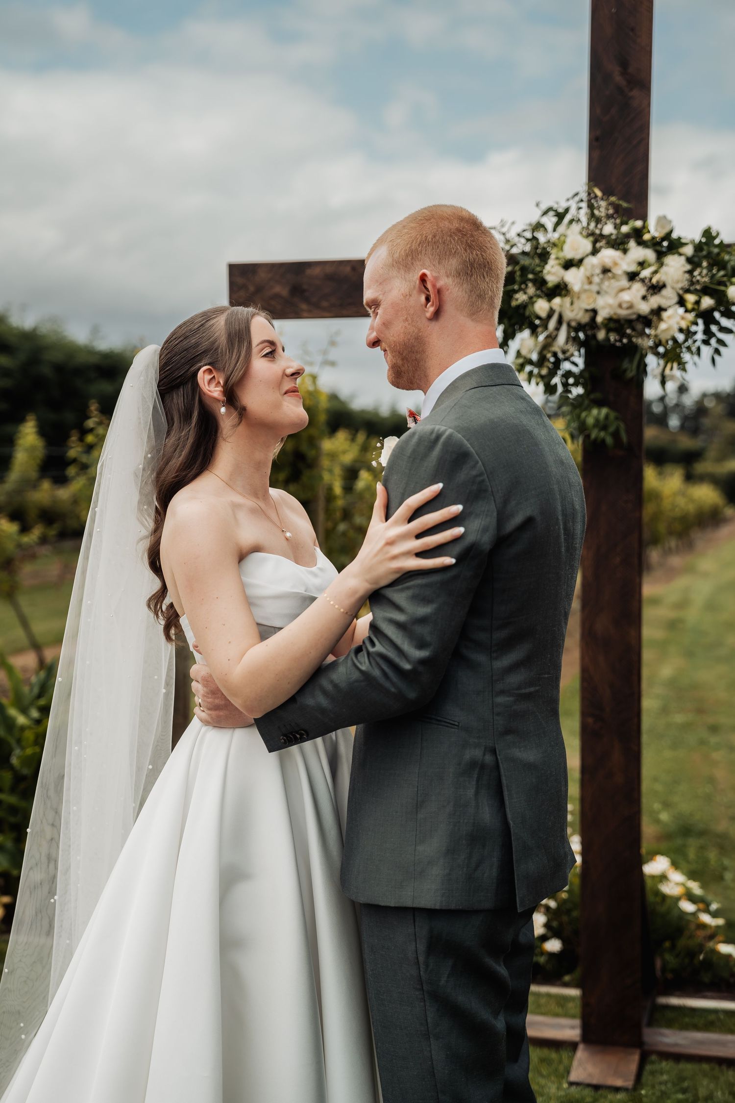 Bride and groom embrace tenderly at outdoor altar decorated with white floral arrangement on wooden cross.