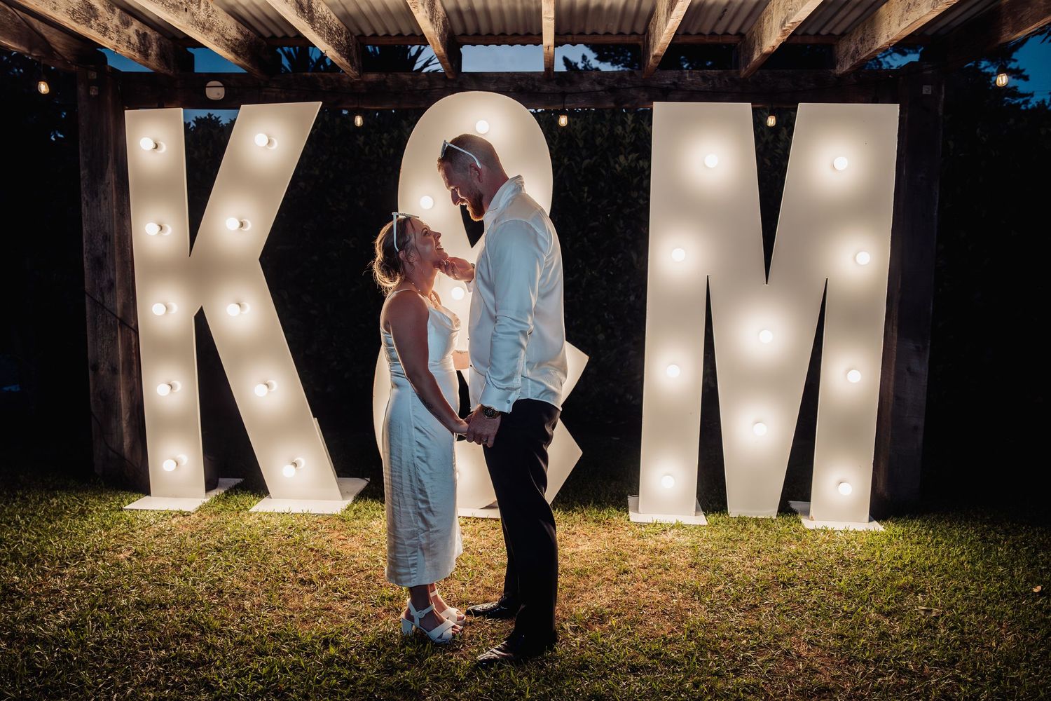 Bride and groom sharing a romantic kiss in front of illuminated marquee letters K and M at night.
