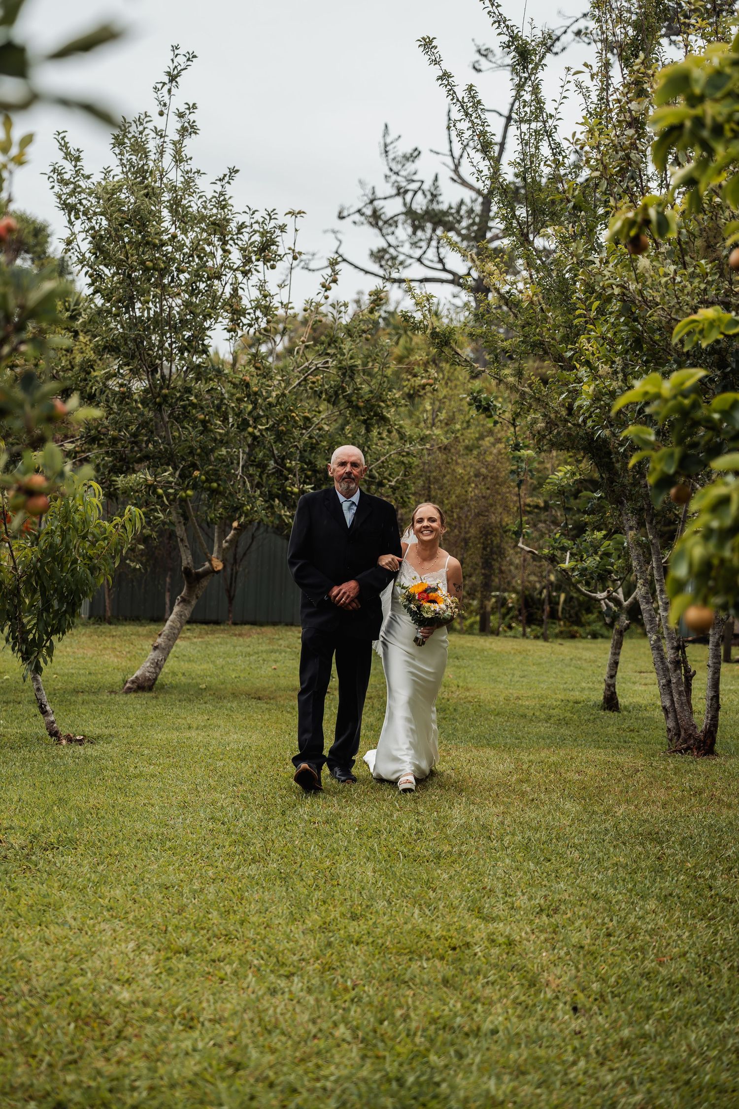 Smiling bride holding bouquet walks with father through apple orchard on wedding day.