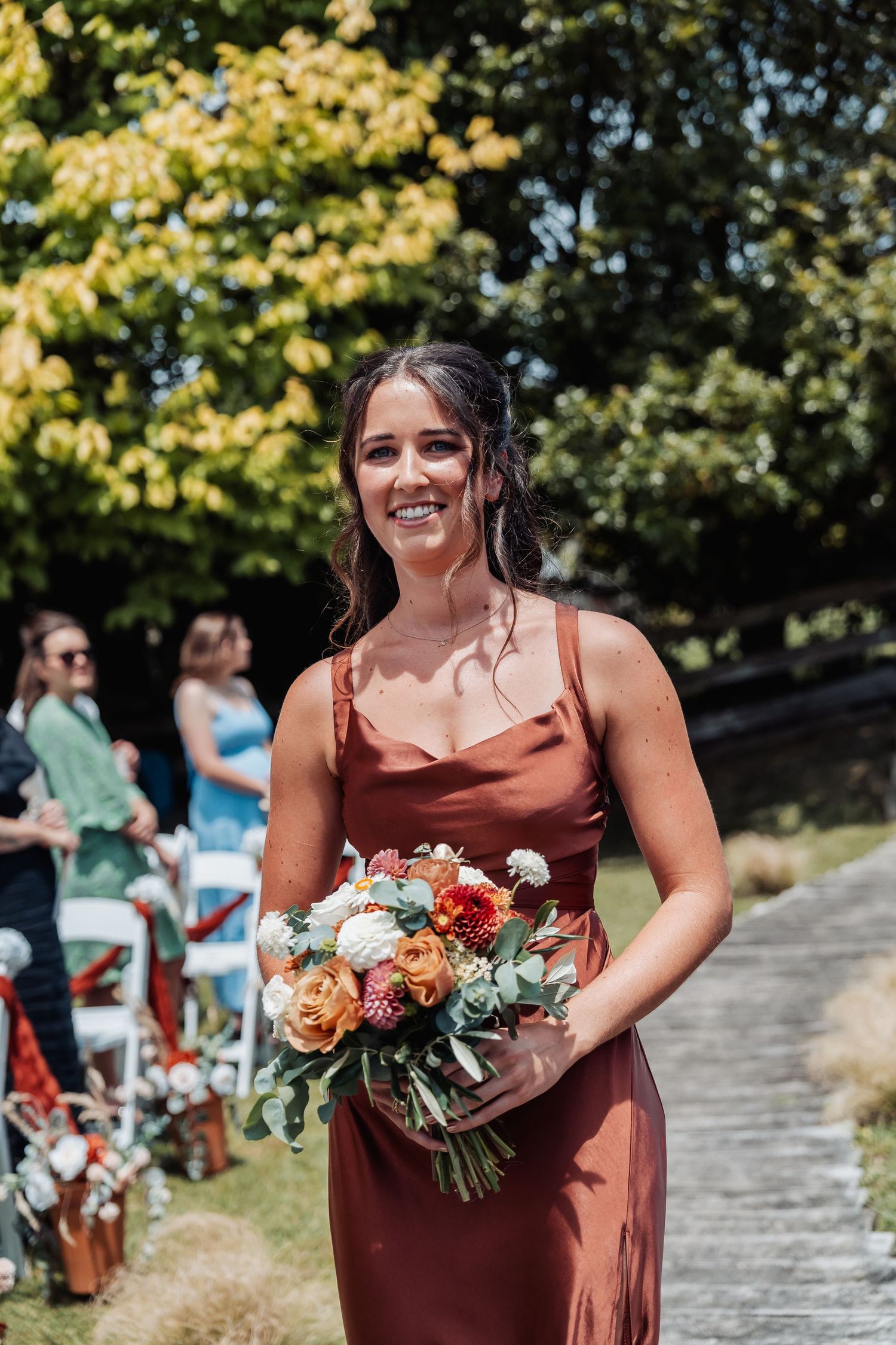 Smiling bridesmaid in rust satin gown walks down the aisle carrying an orange and white bouquet.