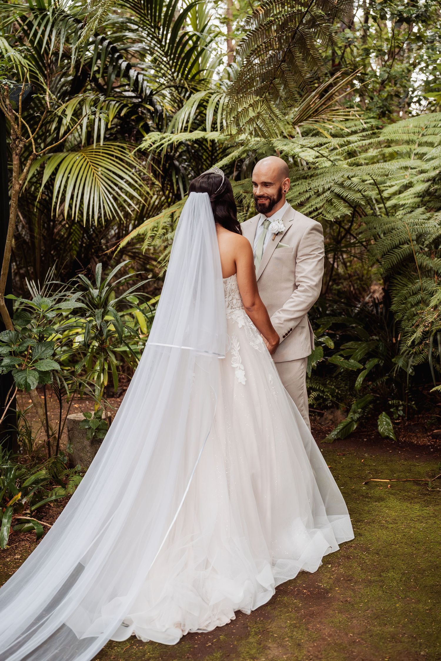 Bride in white gown with long veil holding hands with bald groom in beige suit in lush garden.