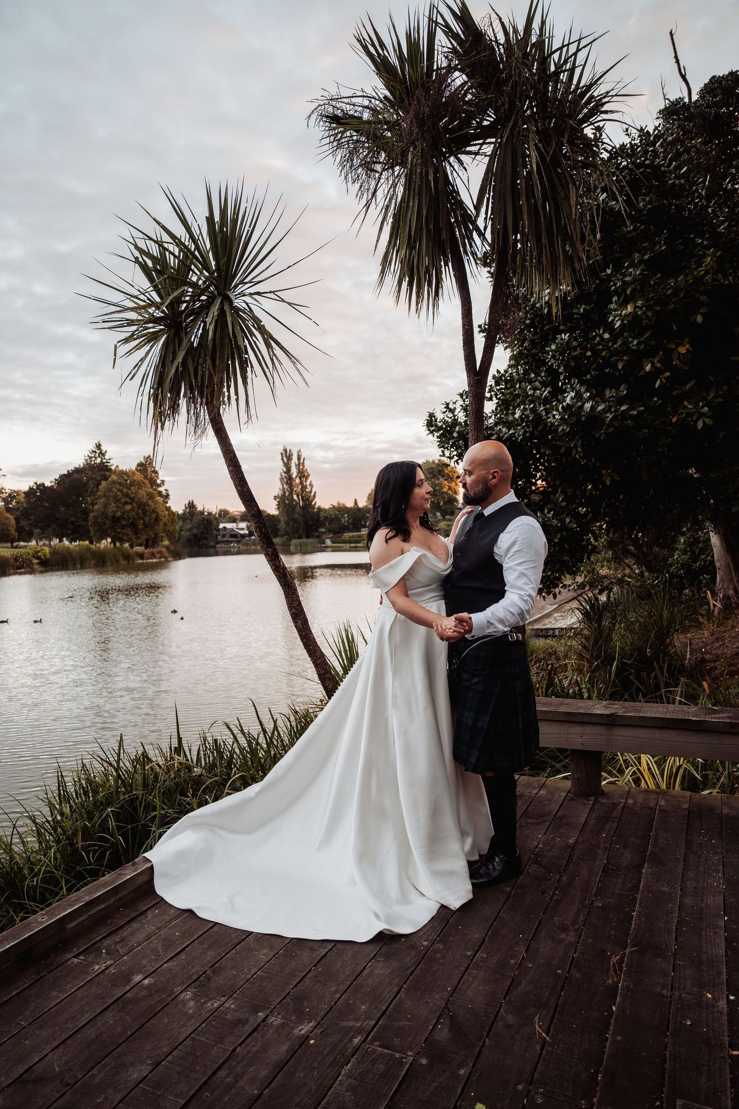 Bride and groom embrace near lakeside at dusk with tall palm trees and dramatic cloudy sky behind them.