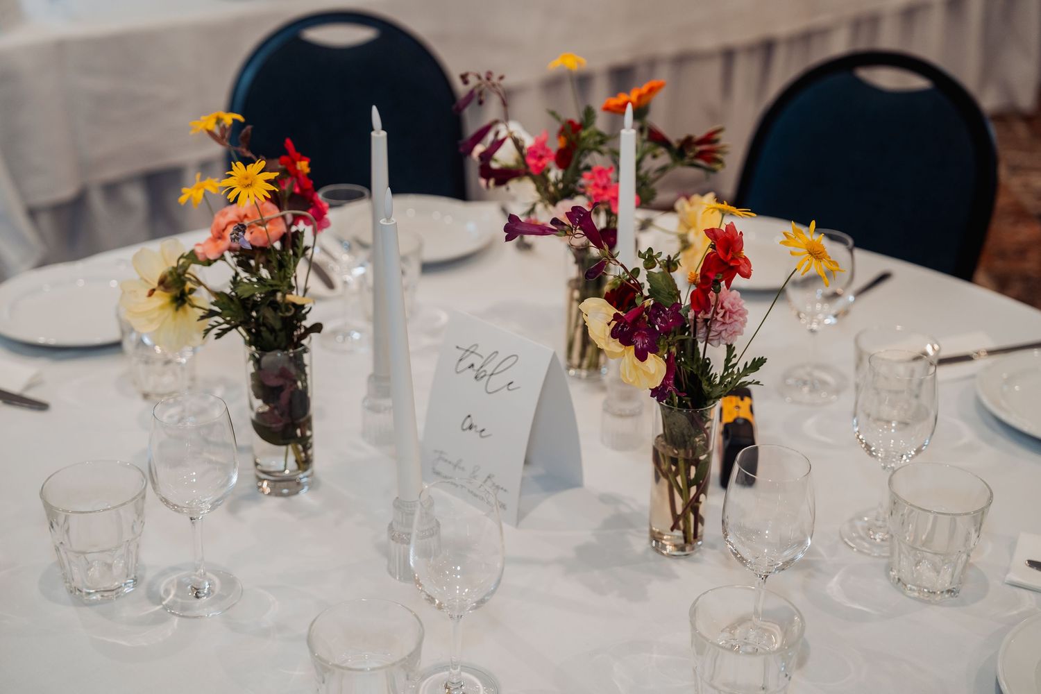 Elegant wedding reception table with colorful wildflower arrangements, white taper candle, crystal glasses, and a table number card.