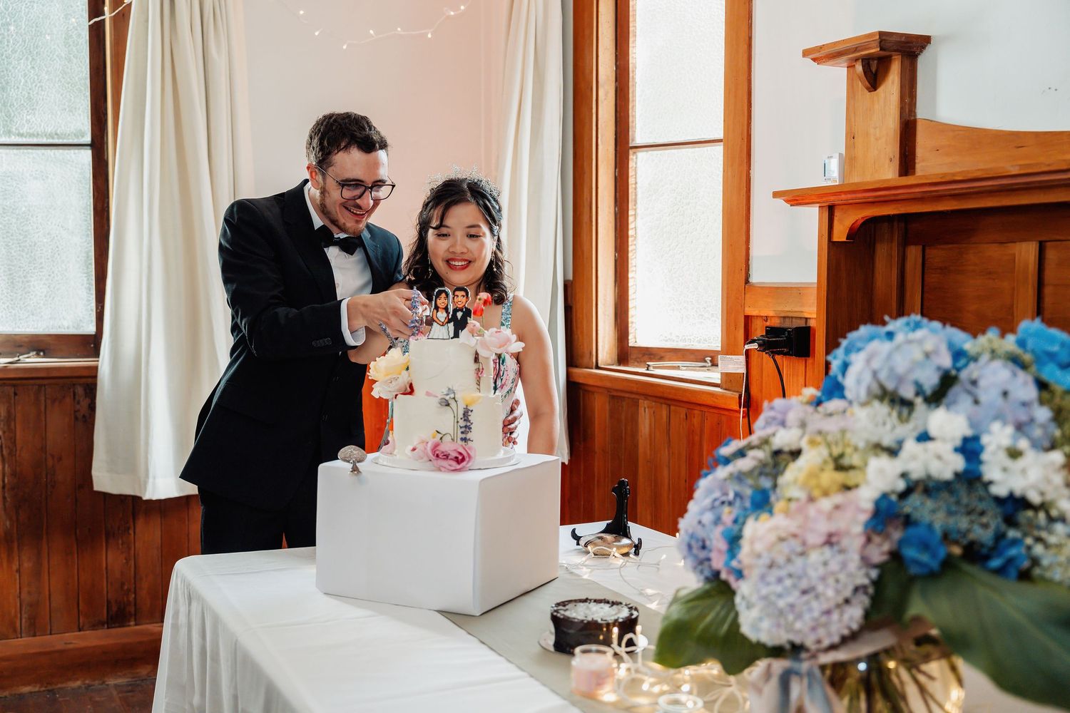 Newlyweds cut white wedding cake together indoors with blue hydrangea floral centerpiece nearby.