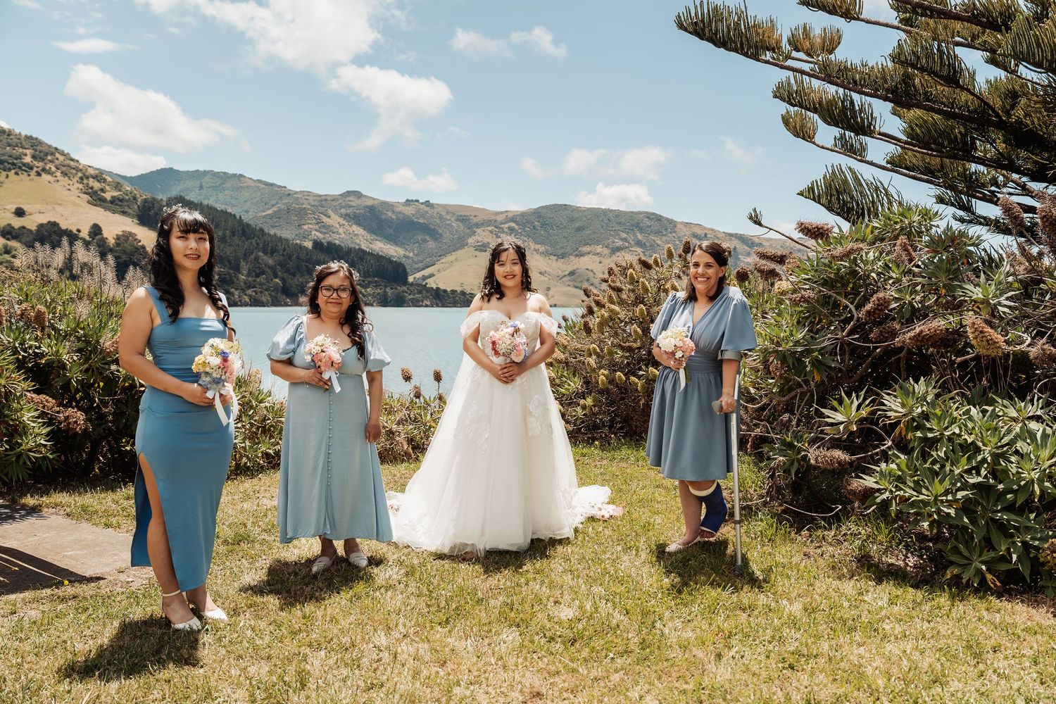 Bride in white gown with three bridesmaids in blue dresses posing outdoors with mountains and lake backdrop.