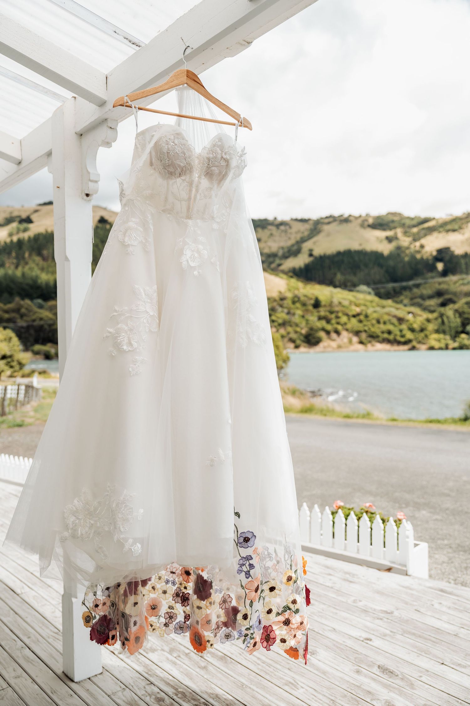 White wedding dress with floral embroidery hem hanging on wooden hanger at scenic waterfront venue.