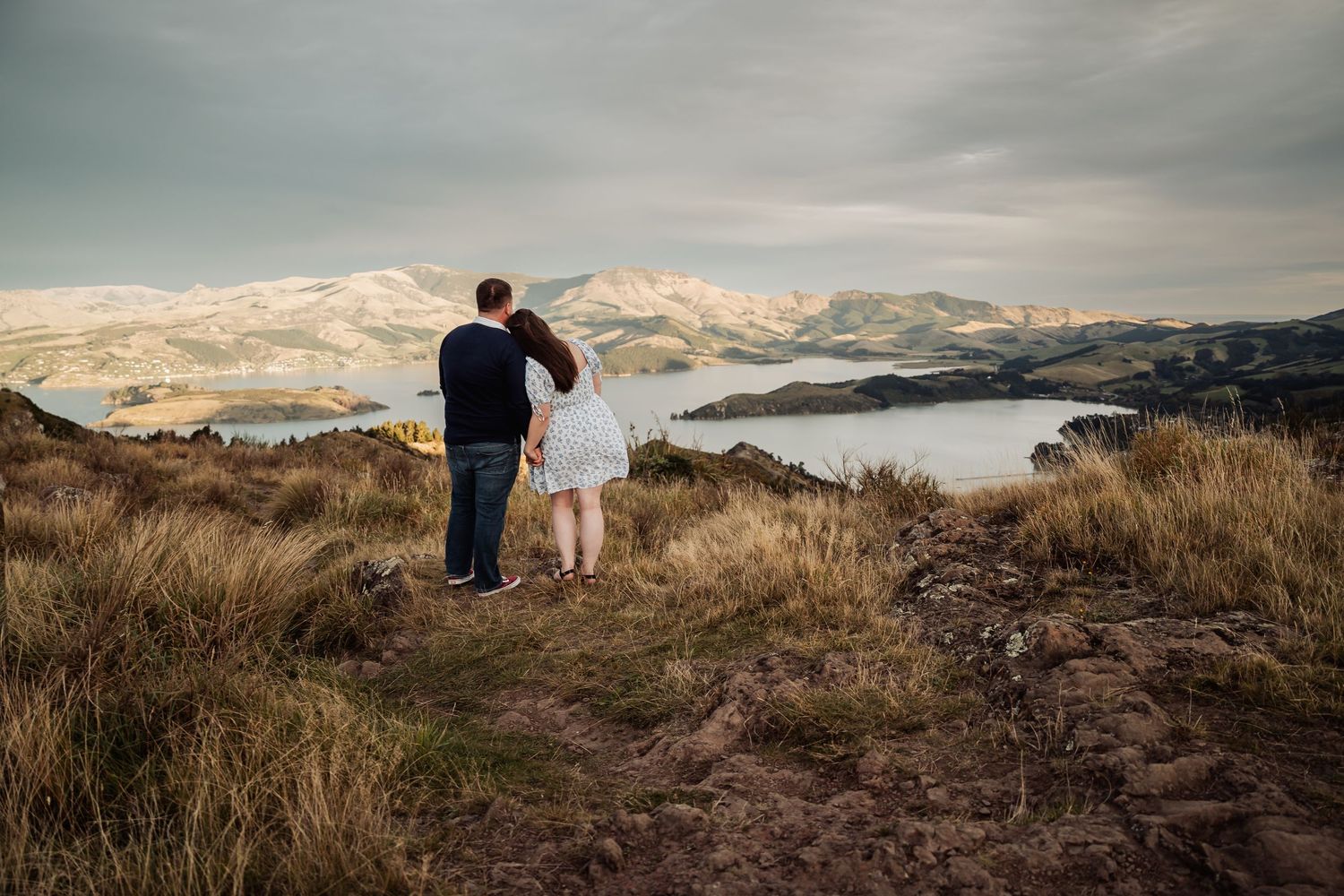 Couple embracing on a hilltop overlooking a scenic bay with snow-capped mountains under a cloudy sky.