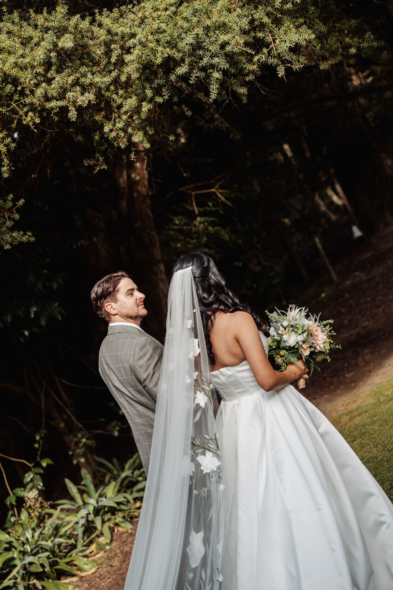 Bride and groom walk through lush garden, bride holding bouquet with long veil trailing.