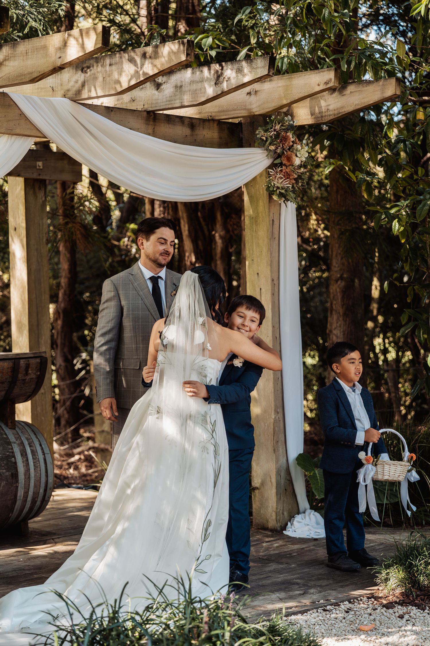Young boy hugs bride at rustic outdoor wedding ceremony under wooden pergola with white draping.