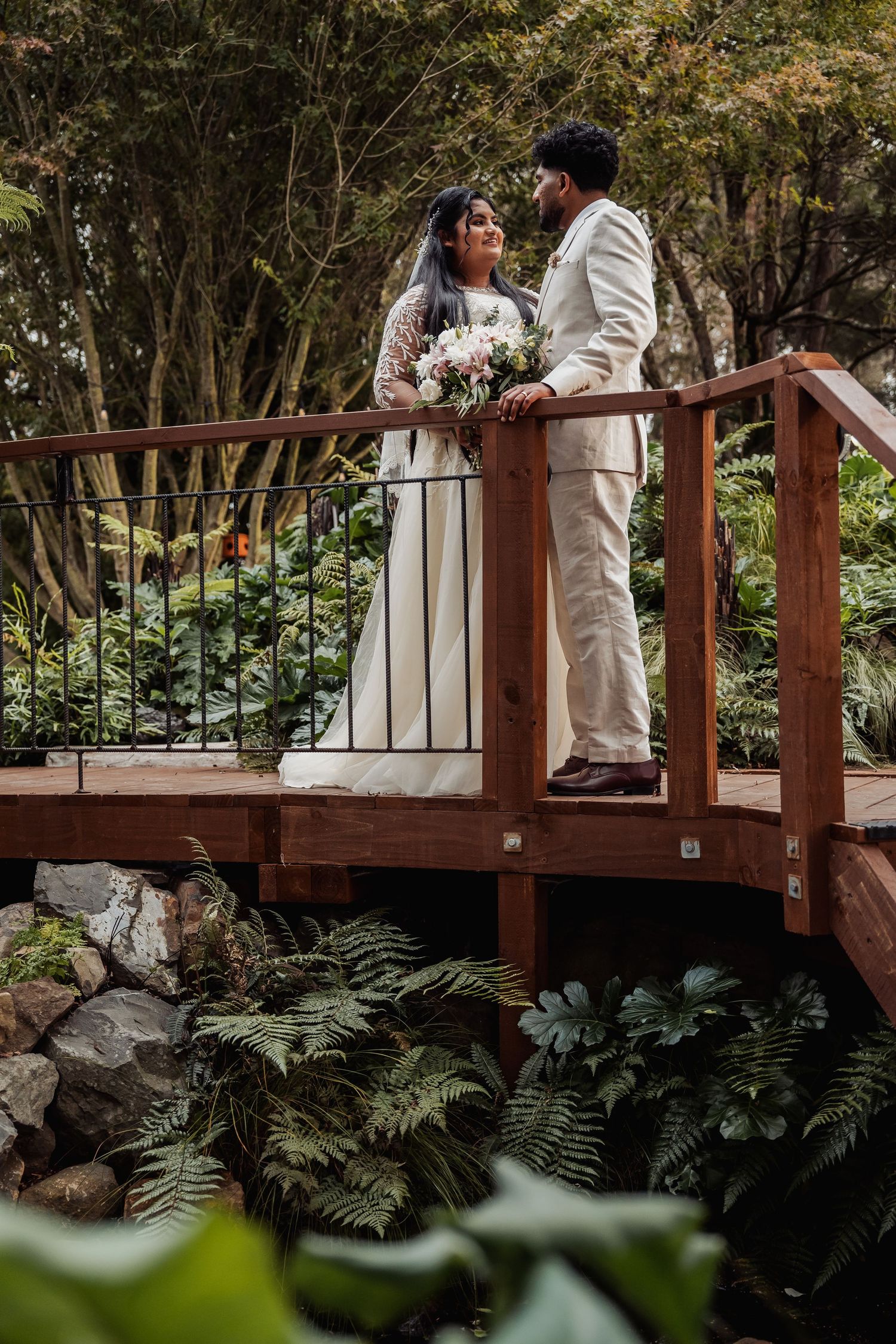 Bride and groom share a romantic moment on a wooden bridge surrounded by lush green garden foliage.