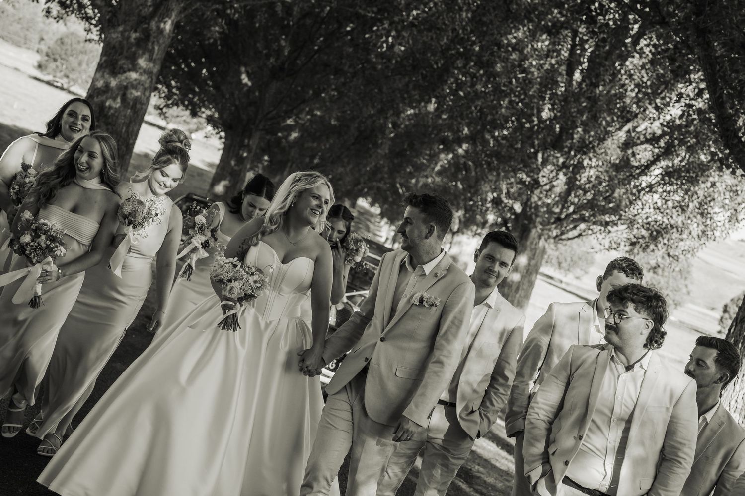 Black and white wedding party photo of bride, groom, bridesmaids and groomsmen under shady trees.