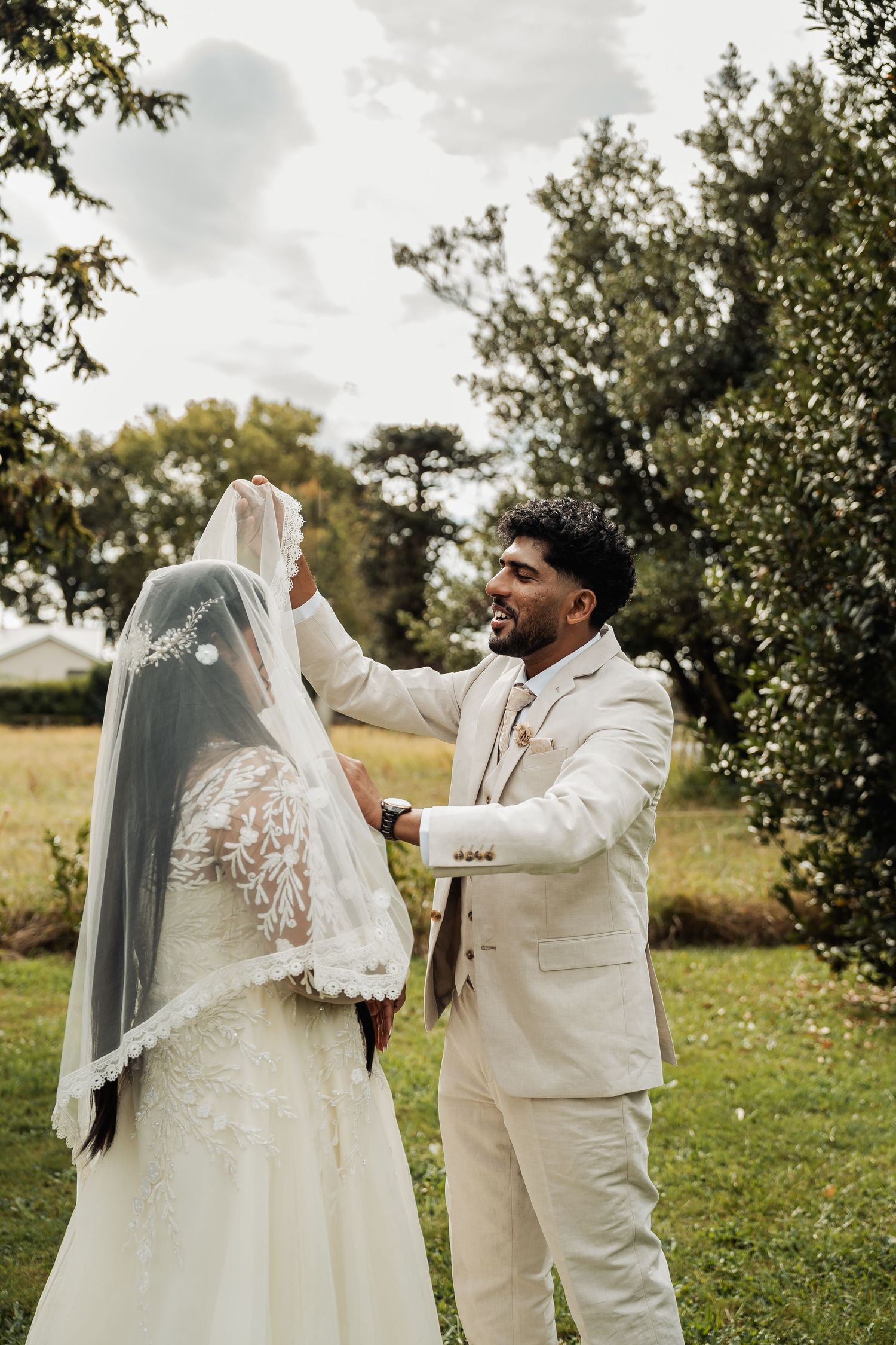 Groom lifting bride's veil in garden setting during outdoor wedding ceremony.