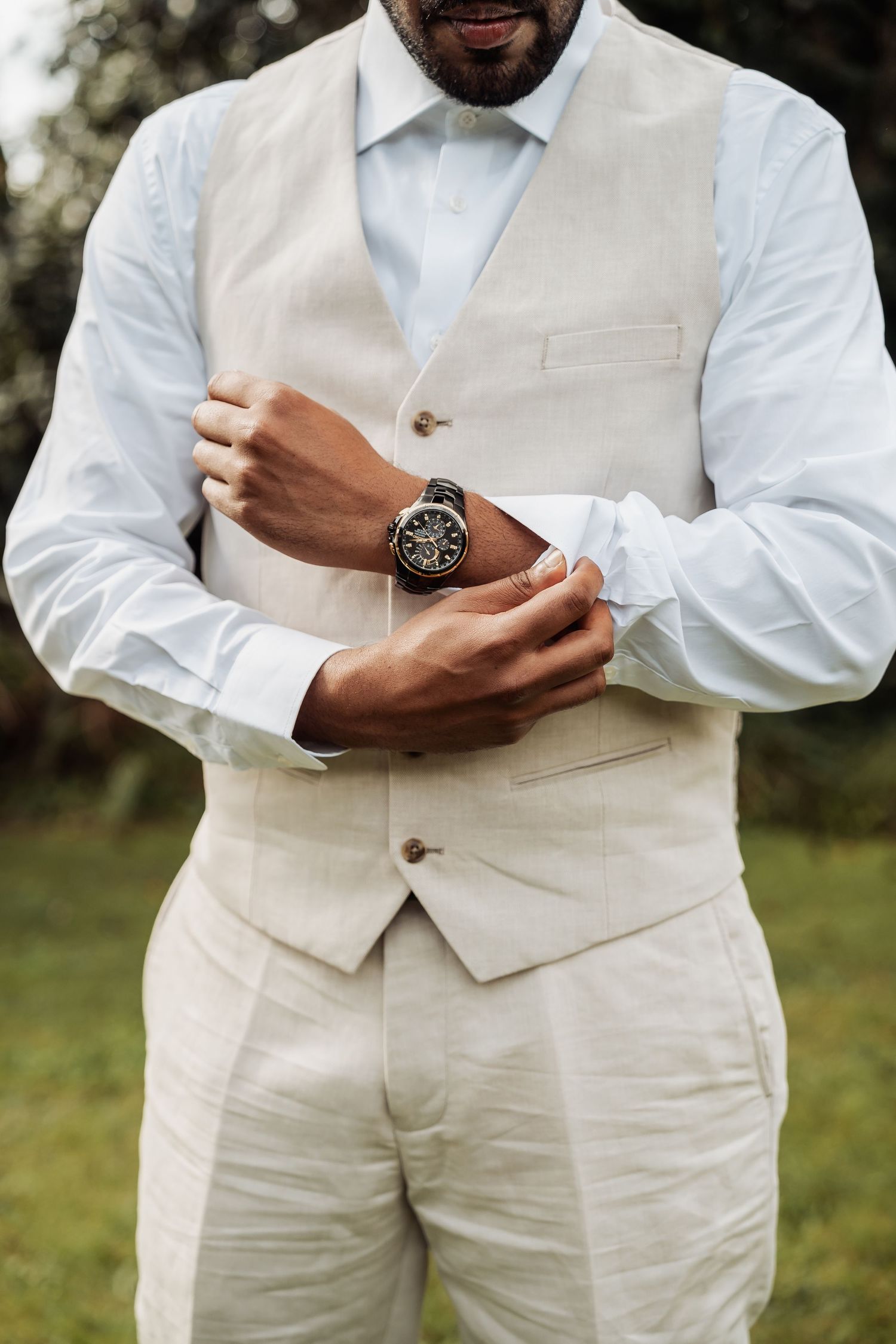 A groom in a beige linen vest and white shirt adjusts his cufflinks while wearing a classic timepiece watch.