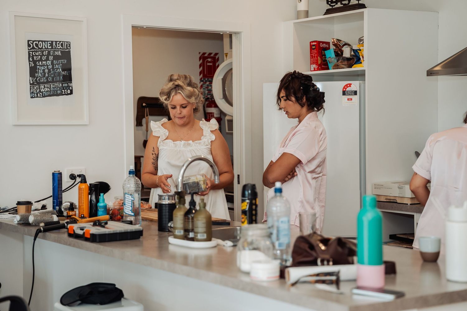 Women preparing for a wedding in a bright kitchen with makeup and hair tools spread across the counter.