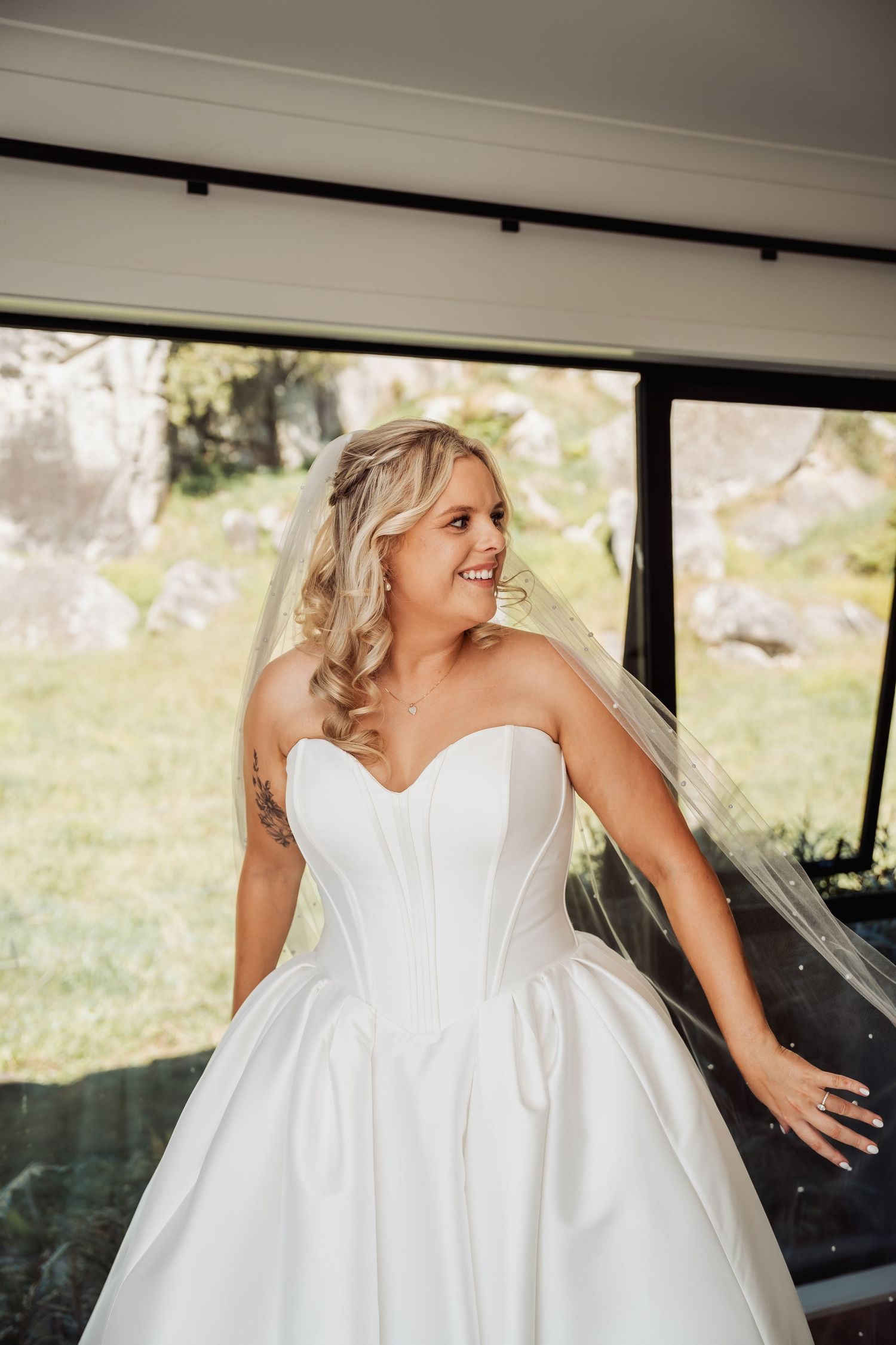 A smiling bride in a strapless white corset ballgown and veil poses by large windows on her wedding day.