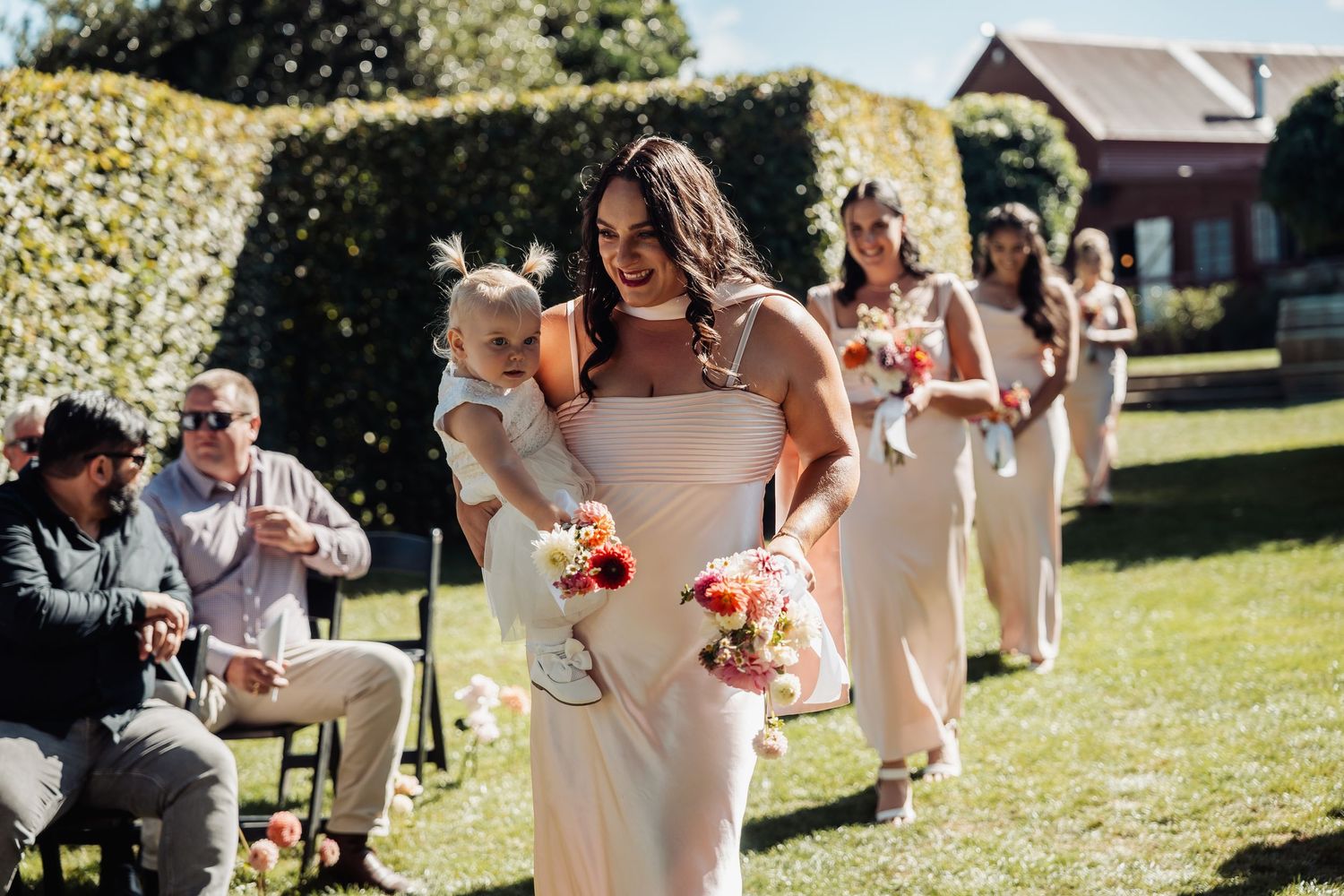 Bridesmaid carrying baby walking down outdoor aisle with bouquet at sunny garden wedding ceremony.